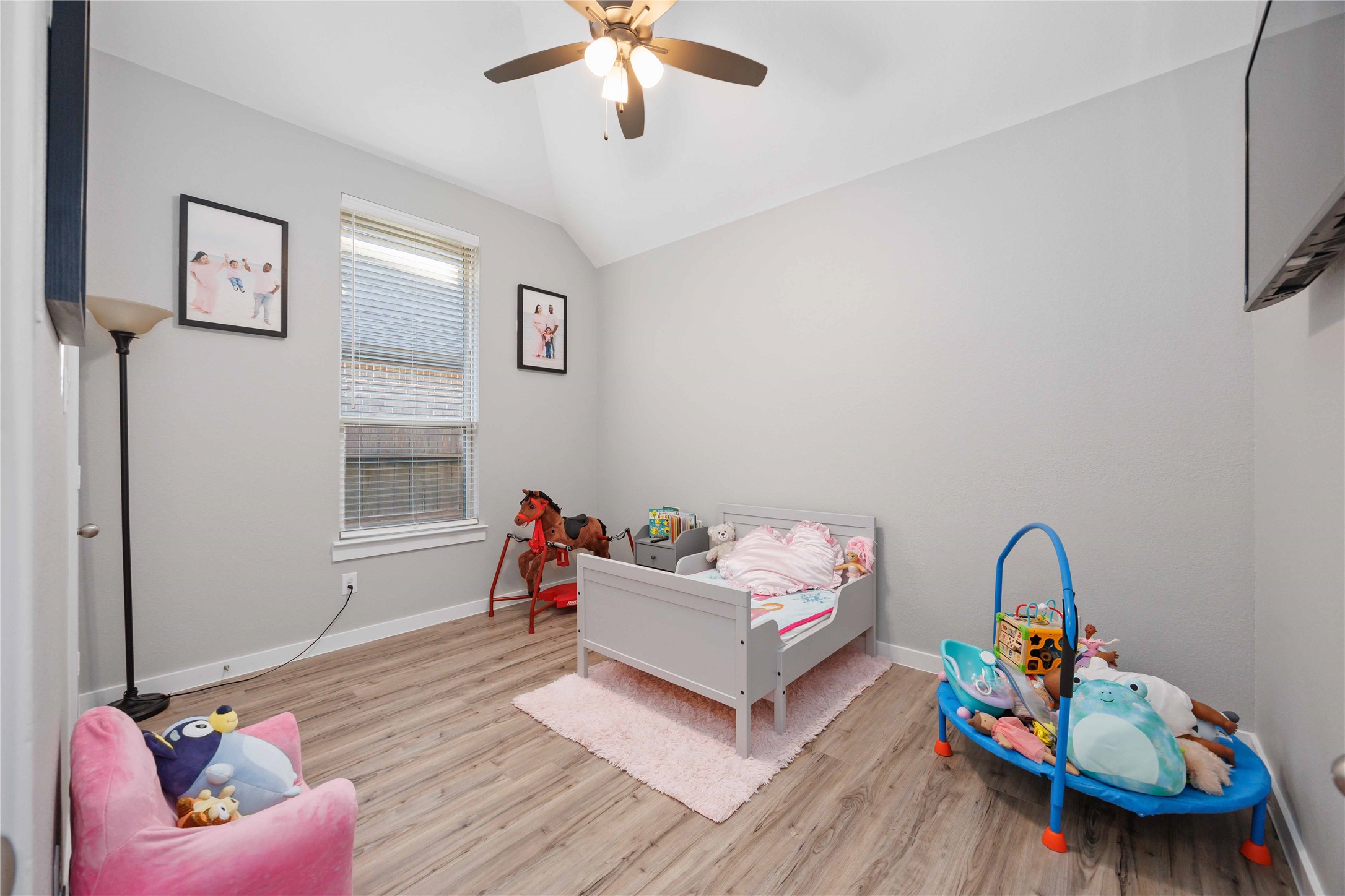 This cozy child's bedroom features a light gray color scheme with wood-look flooring. It includes a small bed, plush toys, a floor lamp, and wall art. The room is well-lit with natural light from a window, complemented by a ceiling fan with lights.