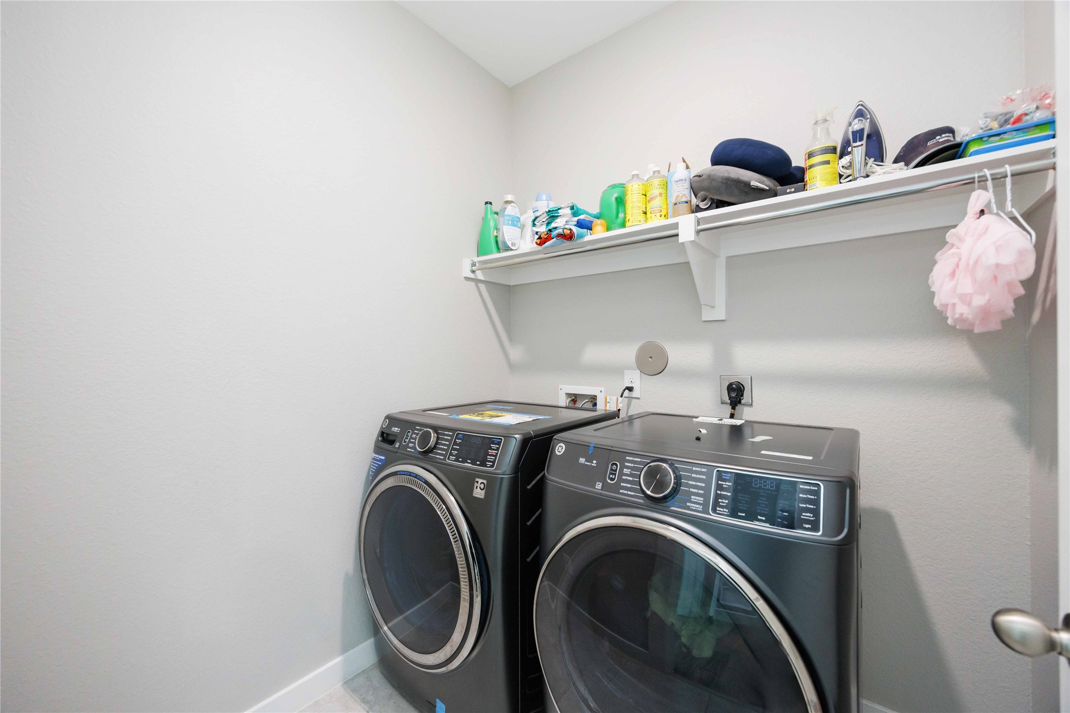 This laundry room features modern black front-loading washer and dryer units. It has a wall-mounted shelf for storage, ideal for organizing cleaning supplies. The space is bright and functional.
