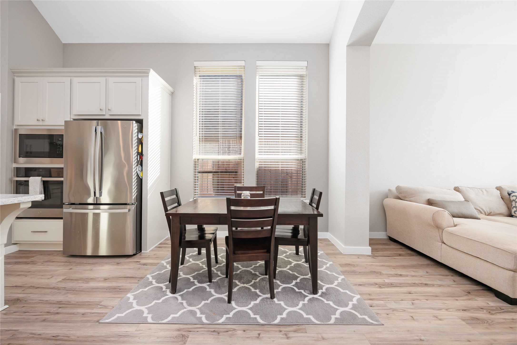 This photo showcases a bright, open kitchen and dining area with modern stainless steel appliances, white cabinetry, and a cozy dining table on a patterned rug. The space flows into a living area with a comfortable beige sofa, all set on warm wood flooring. This photo showcases a bright, open kitchen and dining area with modern stainless steel appliances, white cabinetry, and a cozy dining table on a patterned rug. The space flows into a living area with a comfortable beige sofa, all set on warm wood flooring.