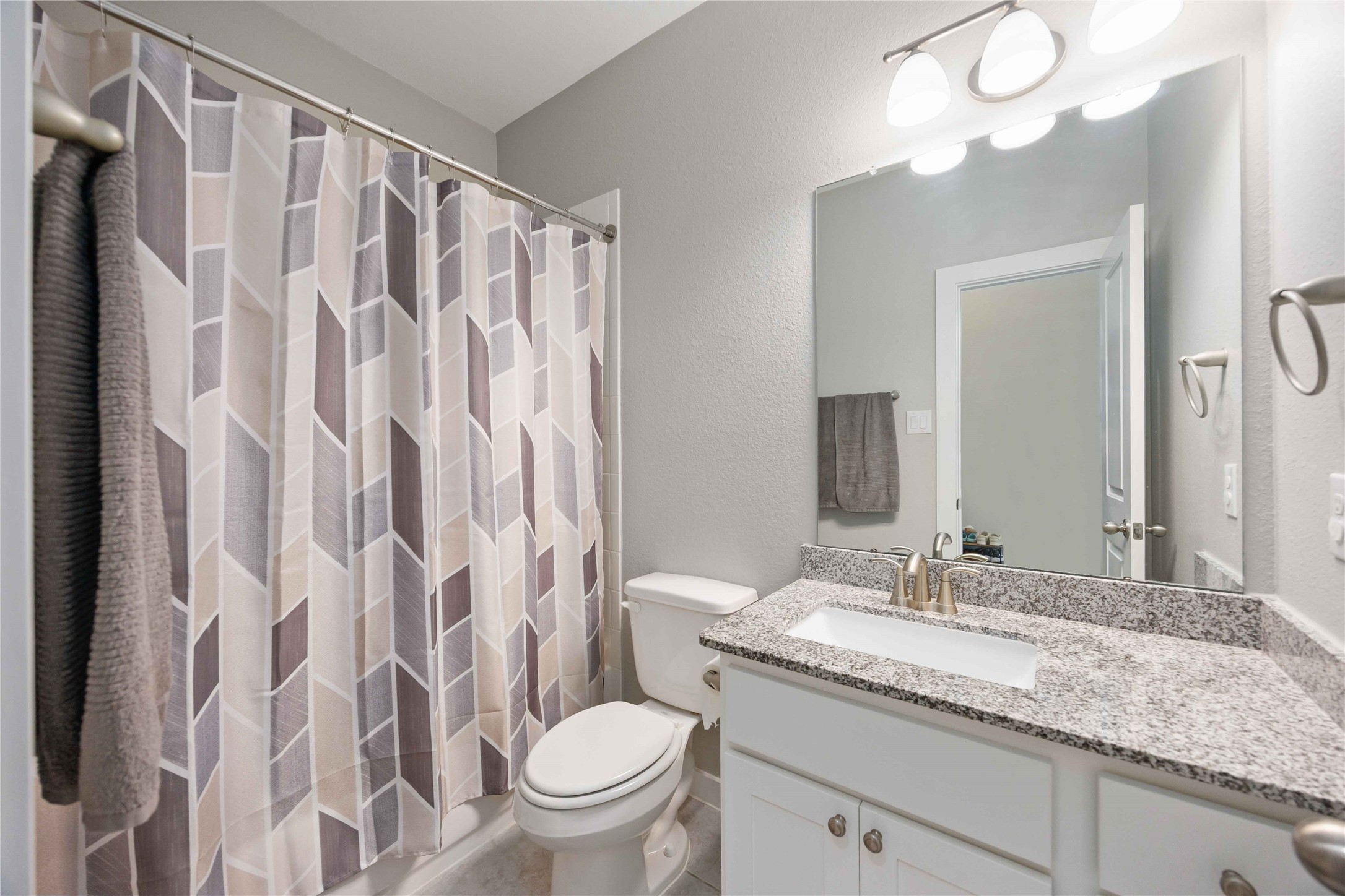 This bathroom features a modern design with a sleek granite countertop, white cabinetry, and a well-lit mirror. It includes a shower with a stylish geometric-patterned curtain, and neutral wall tones create a clean, inviting atmosphere.