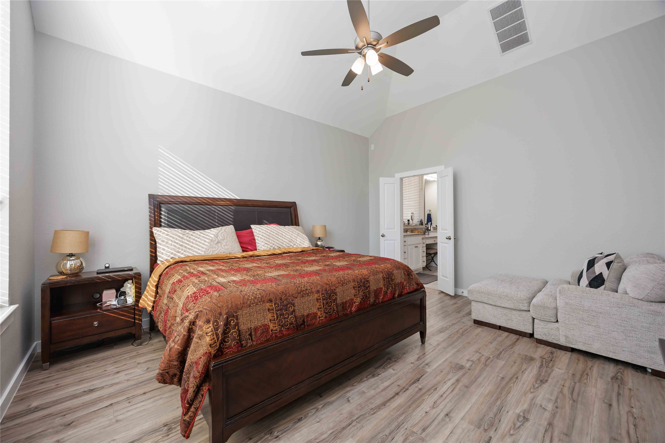 Spacious bedroom with high ceilings, featuring a large bed with elegant bedding, a comfy corner sofa, and an en-suite bathroom. The room has a modern ceiling fan, neutral walls, and plenty of natural light from a nearby window.