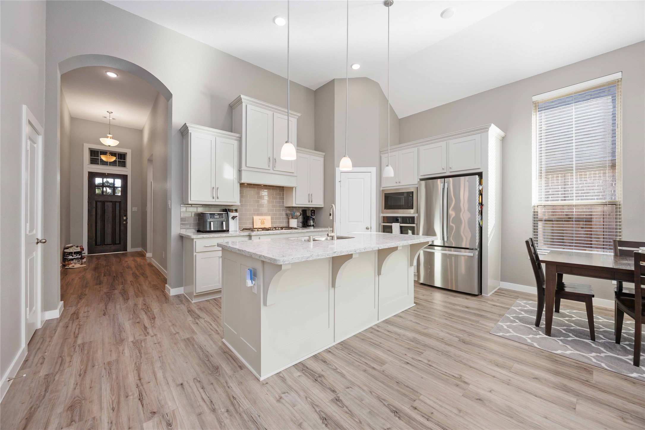 This modern kitchen features high ceilings, sleek stainless steel appliances, and a spacious island with pendant lighting. The open layout includes a dining area next to large windows for natural light. The wood-style flooring adds warmth, and the entryway access provides a welcoming flow. This modern kitchen features high ceilings, sleek stainless steel appliances, and a spacious island with pendant lighting. The open layout includes a dining area next to large windows for natural light. The wood-style flooring adds warmth, and the entryway access provides a welcoming flow.