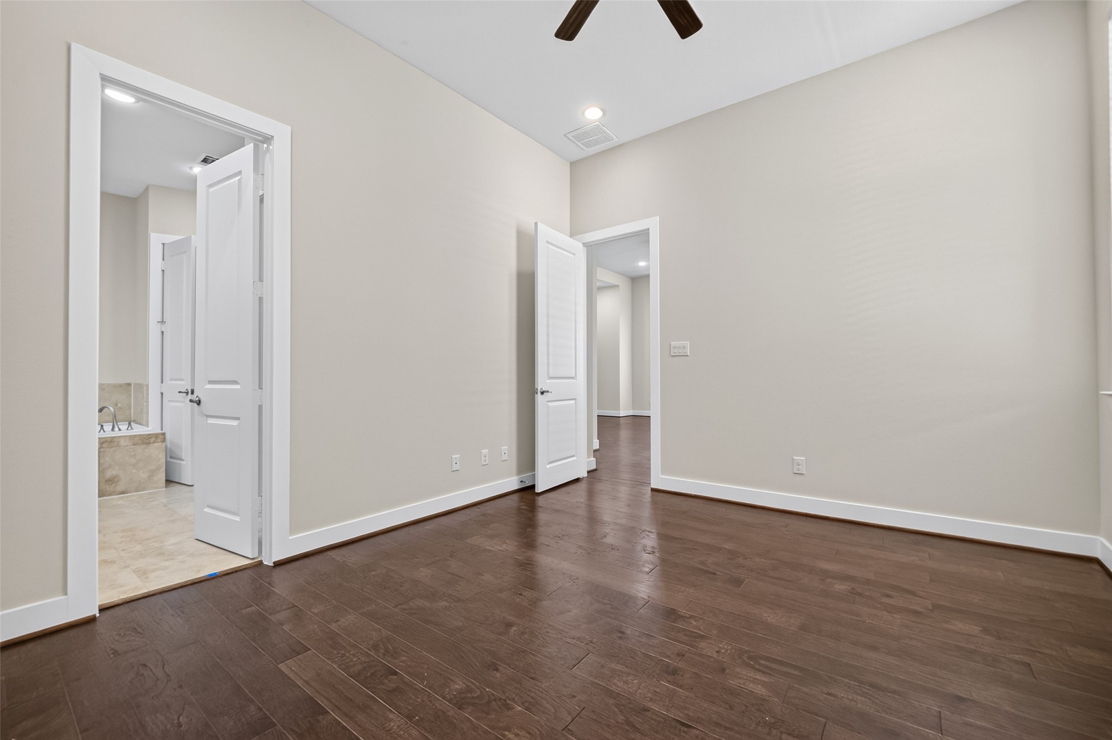 Spacious room with elegant hardwood floors and neutral walls, featuring a ceiling fan and access to a tiled bathroom with a soaking tub.