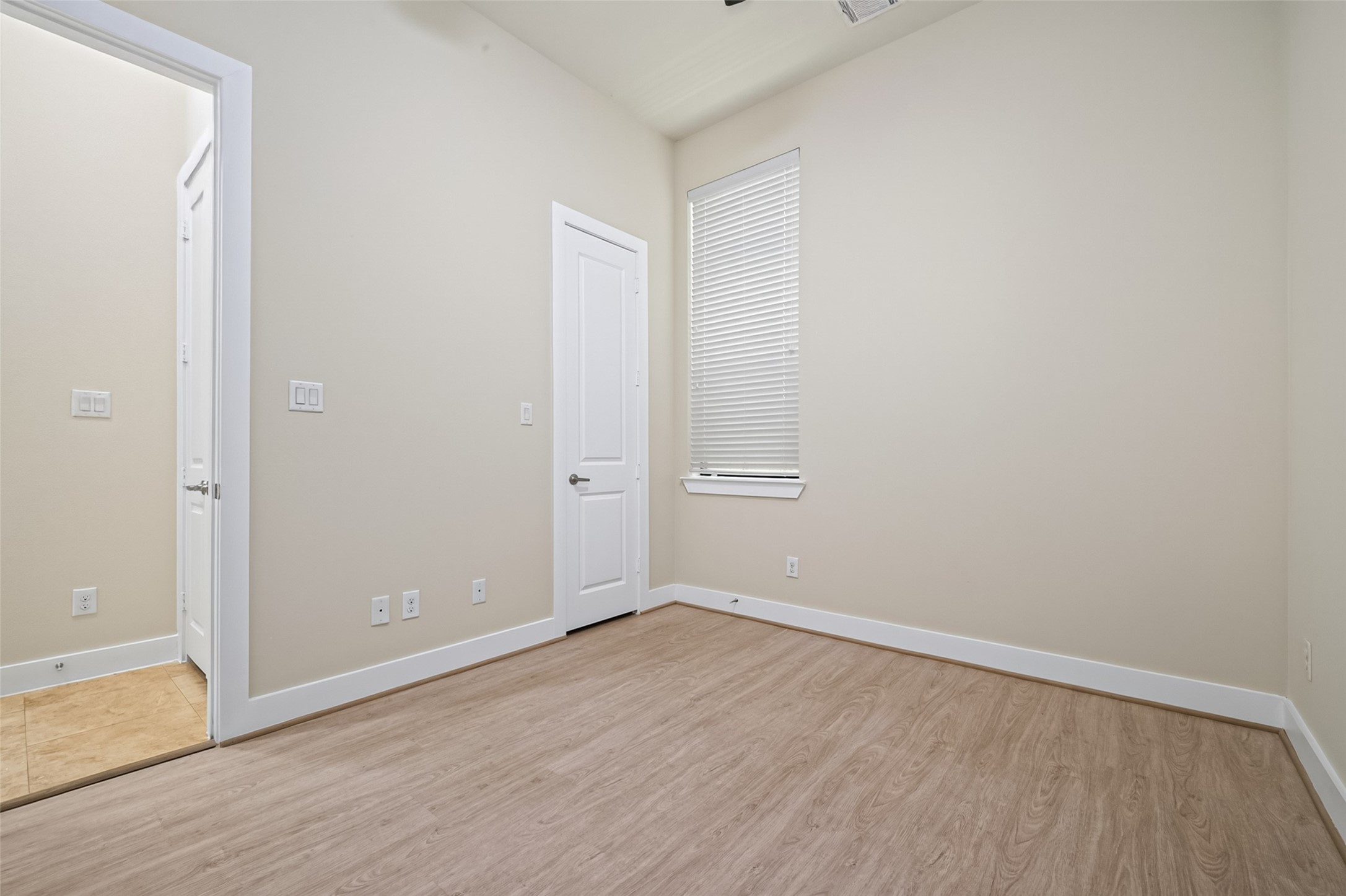 Bright and inviting room with light wood flooring, neutral walls, and ample natural light from a large window, offering a versatile space for a home office or guest bedroom.