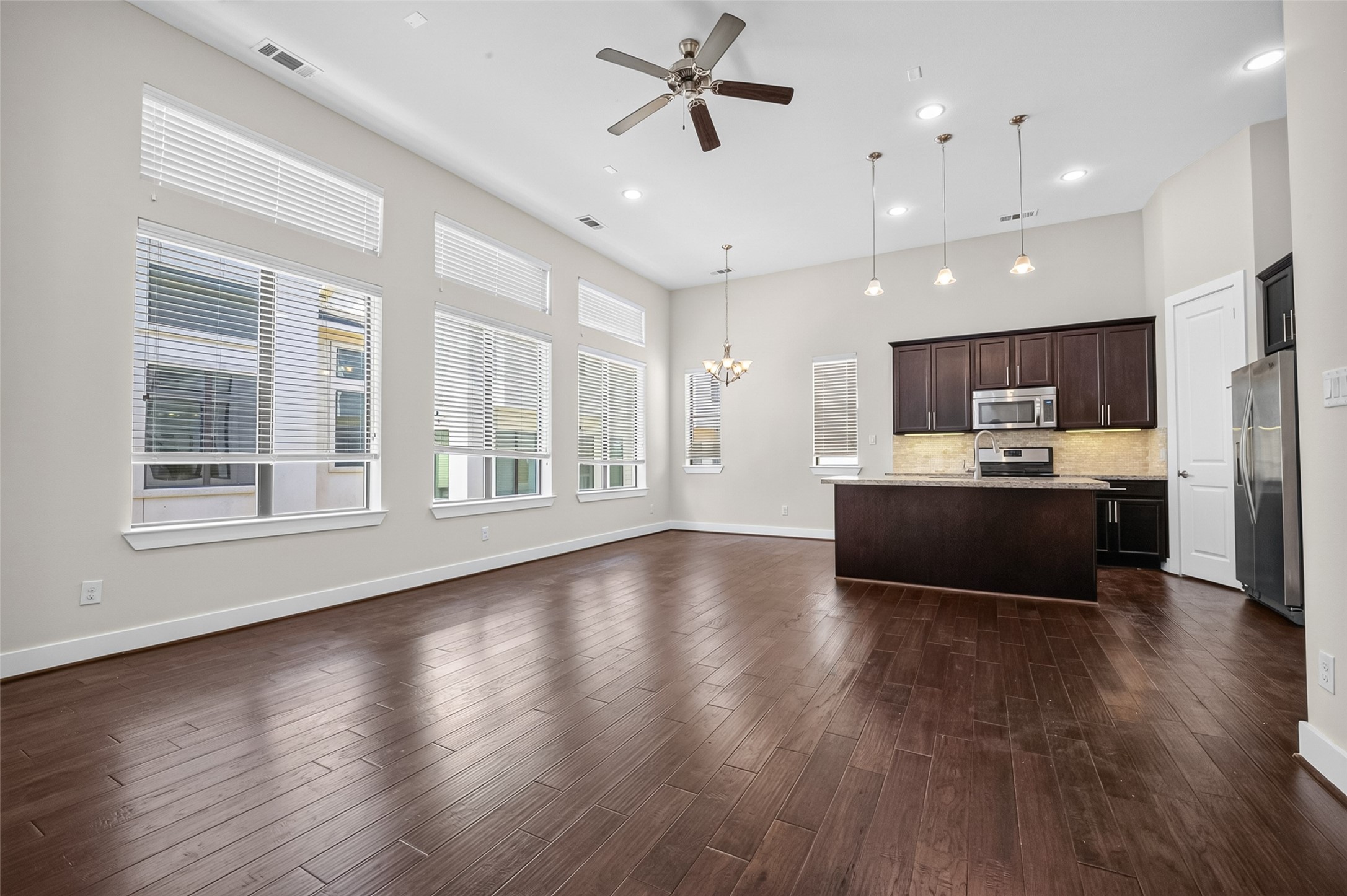 Spacious open-concept living area with high ceilings, abundant natural light from large windows, and a modern kitchen featuring dark cabinetry, stainless steel appliances, and a central island with pendant lighting.