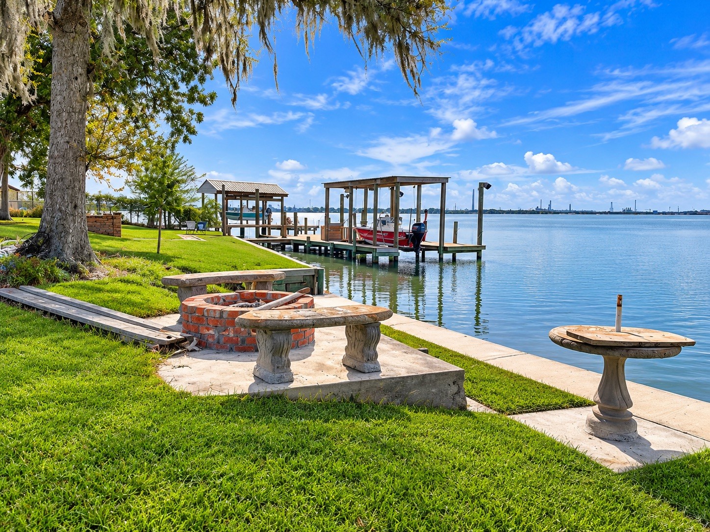 Serene waterfront setting featuring a lush green lawn, charming brick fire pit, and private dock with boat access, perfect for outdoor relaxation and entertainment.