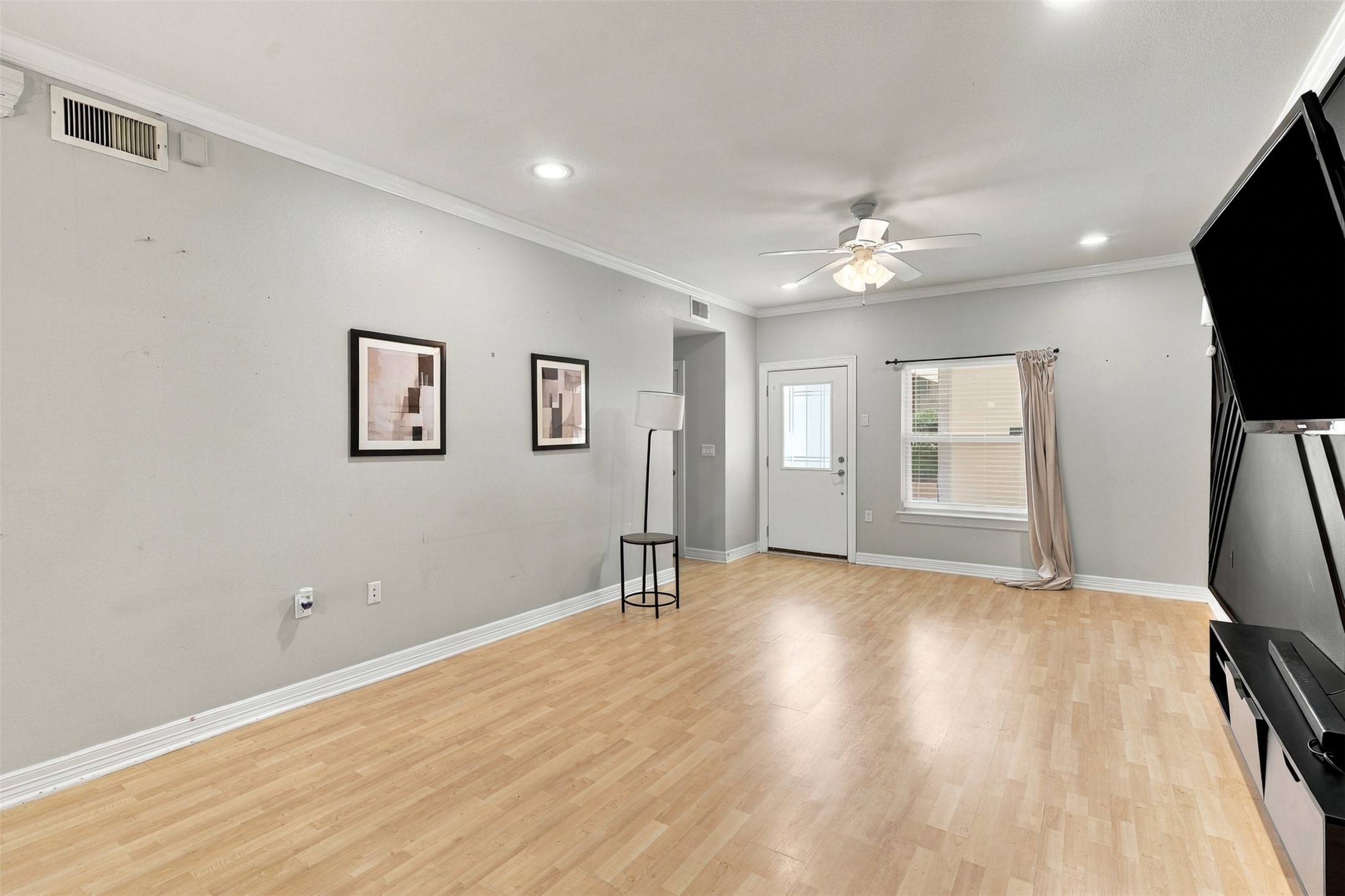 Spacious living area with light wood flooring, neutral walls, and abundant natural light from a large window. The ceiling fan and recessed lighting add modern comfort.