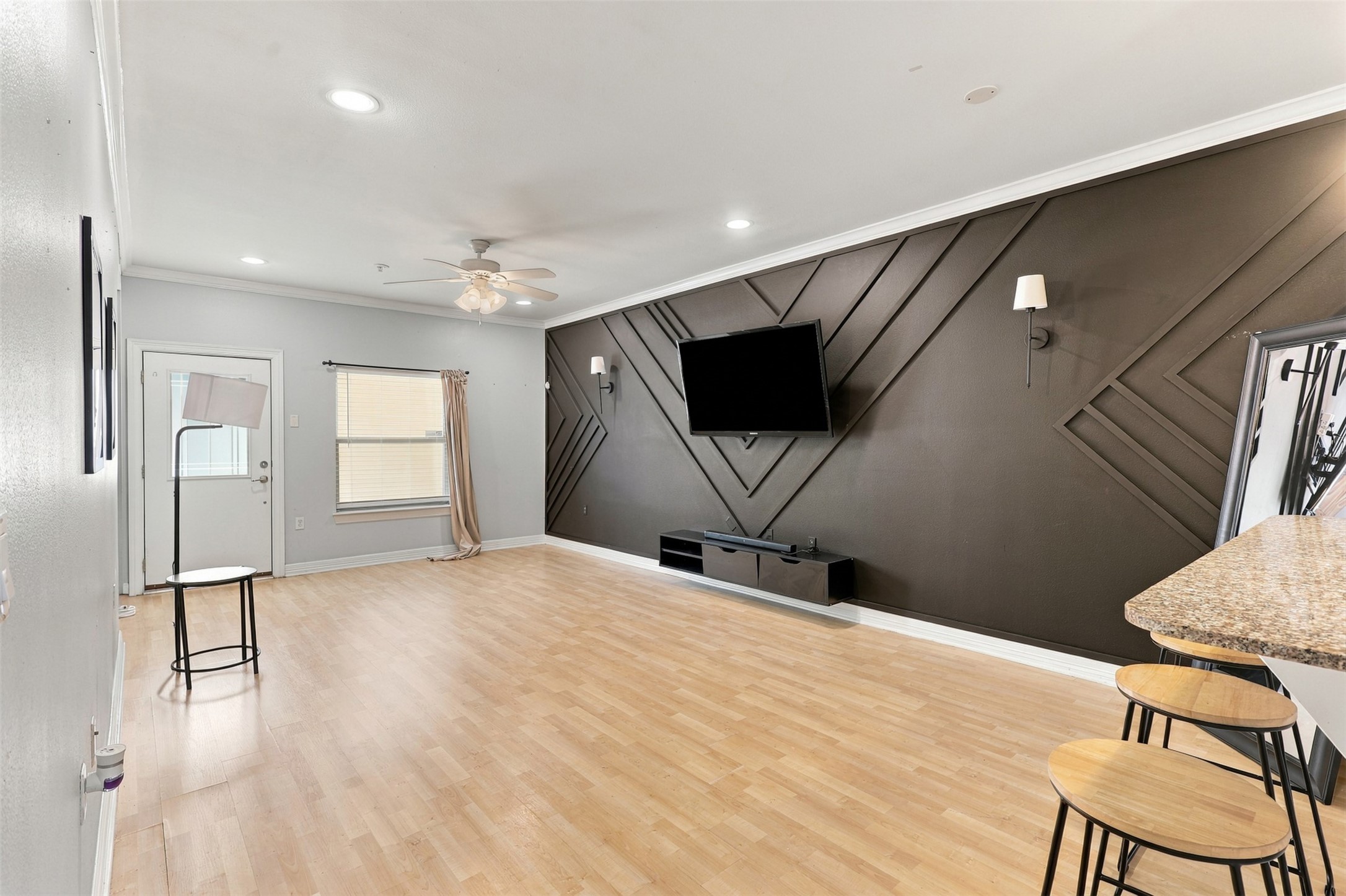 Spacious living area with modern geometric accent wall, featuring a mounted TV and sleek wood flooring. Ample natural light flows through the window, complemented by recessed lighting and a ceiling fan. A granite breakfast bar with stool seating adds a touch of elegance.