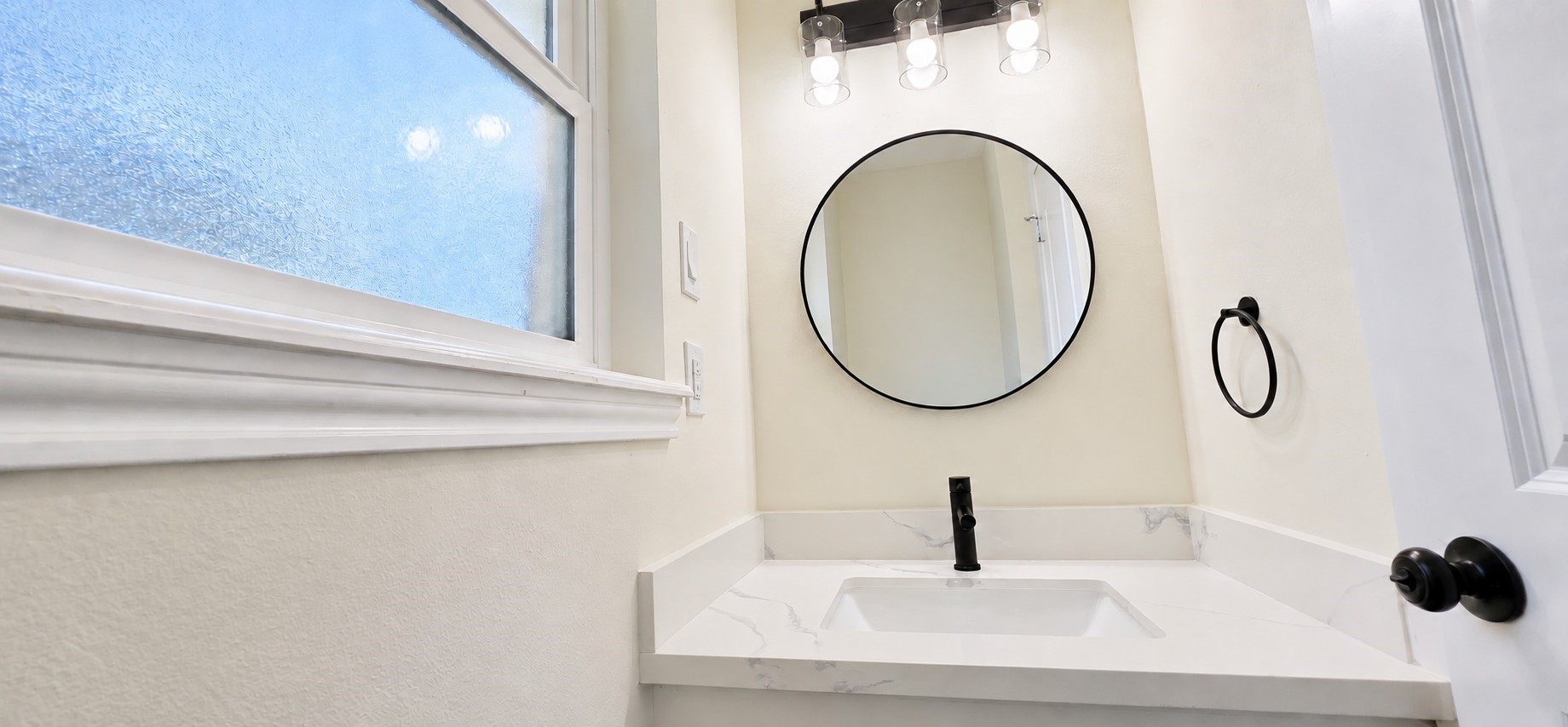 Stylish powder room features a veined countertop, matte-black faucet, round mirror, and frosted window.
