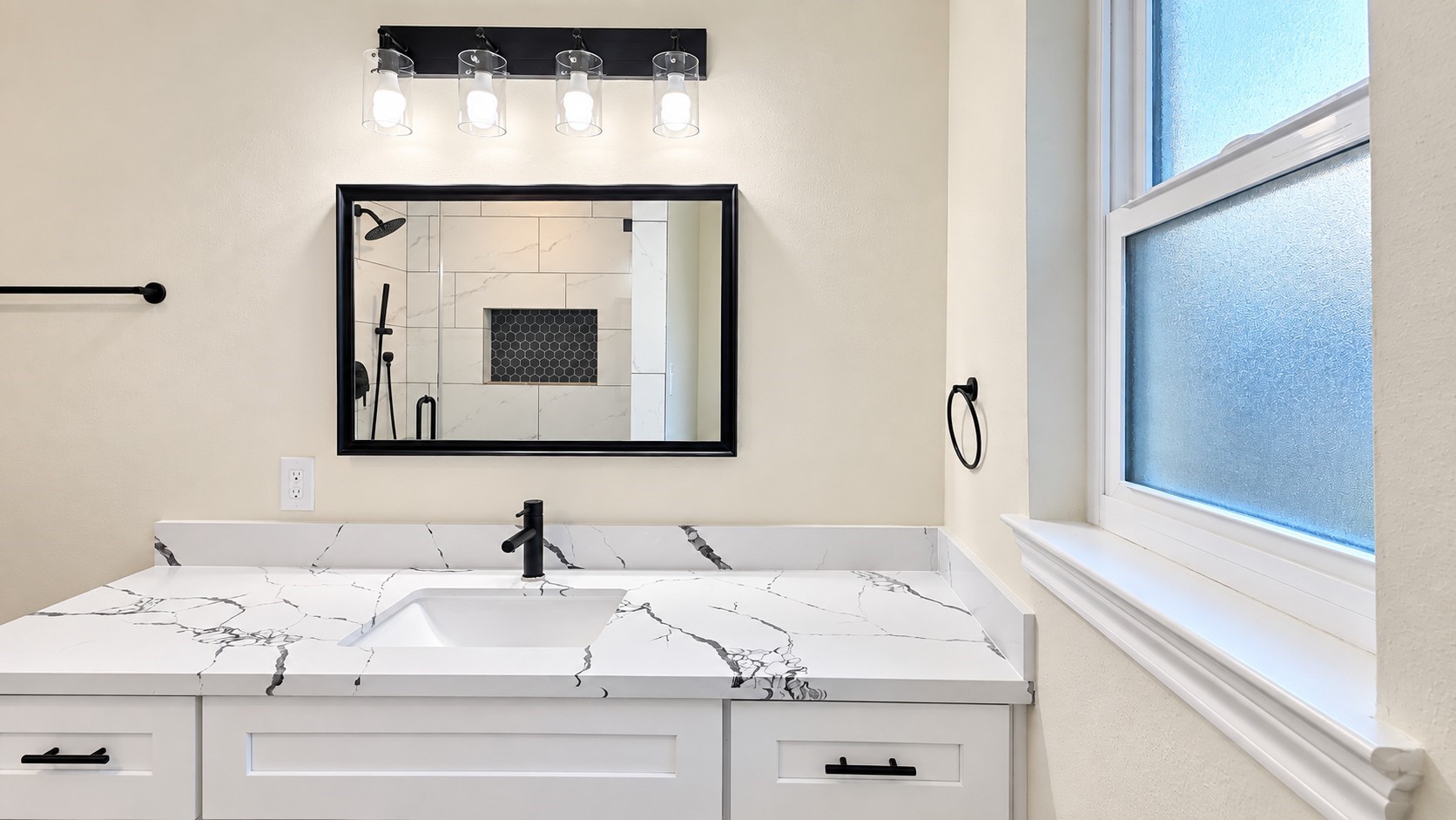Updated primary bath vanity pairs veined countertops, matte-black fixtures, and natural light from the frosted window.