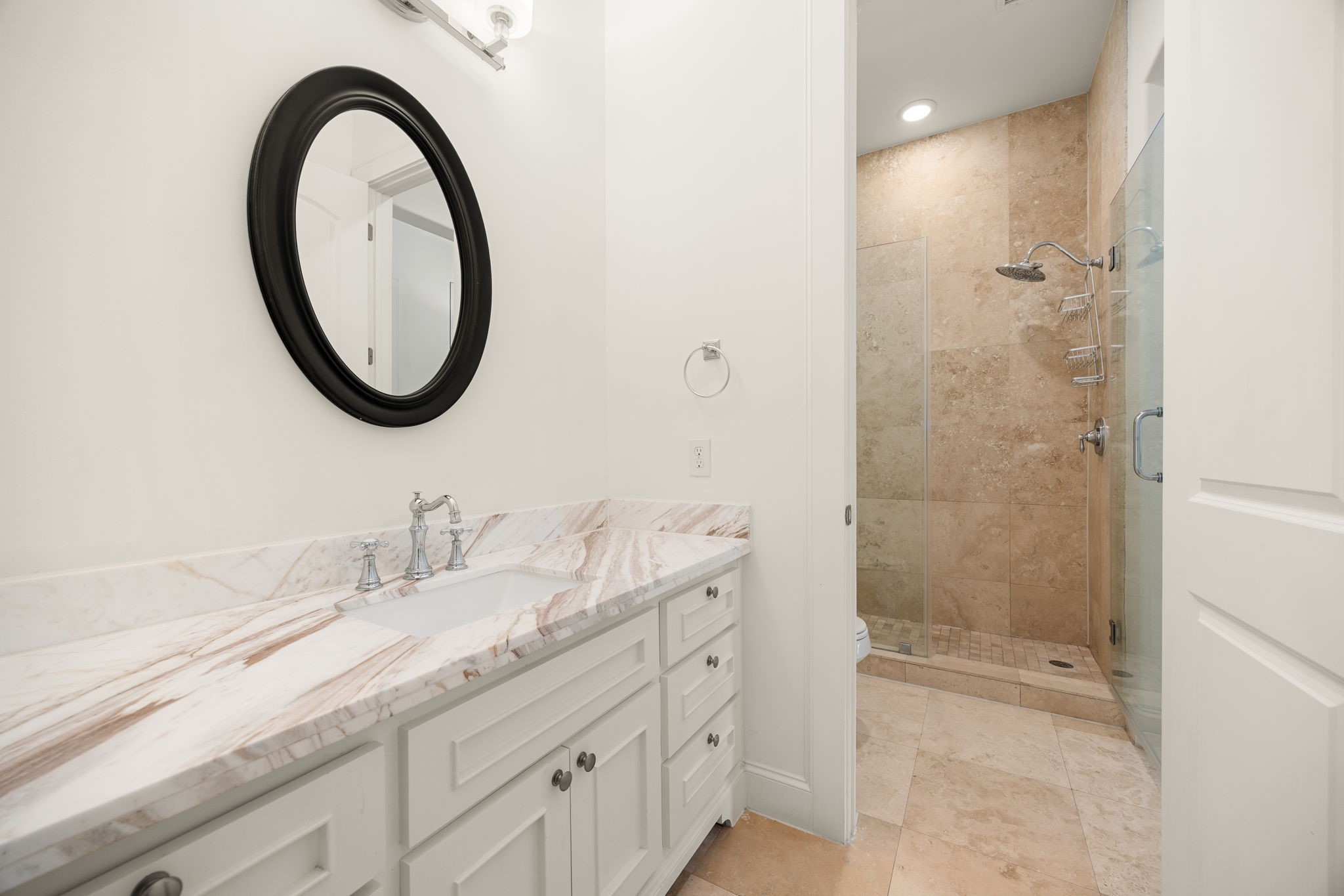 This full bathroom offers a refined blend of comfort and style, featuring crisp white cabinetry, elegant marble countertops, and a spacious walk in shower with glass enclosure and warm stone tile. Thoughtfully finished and beautifully appointed, it provides a welcoming space for guests to refresh and unwind.