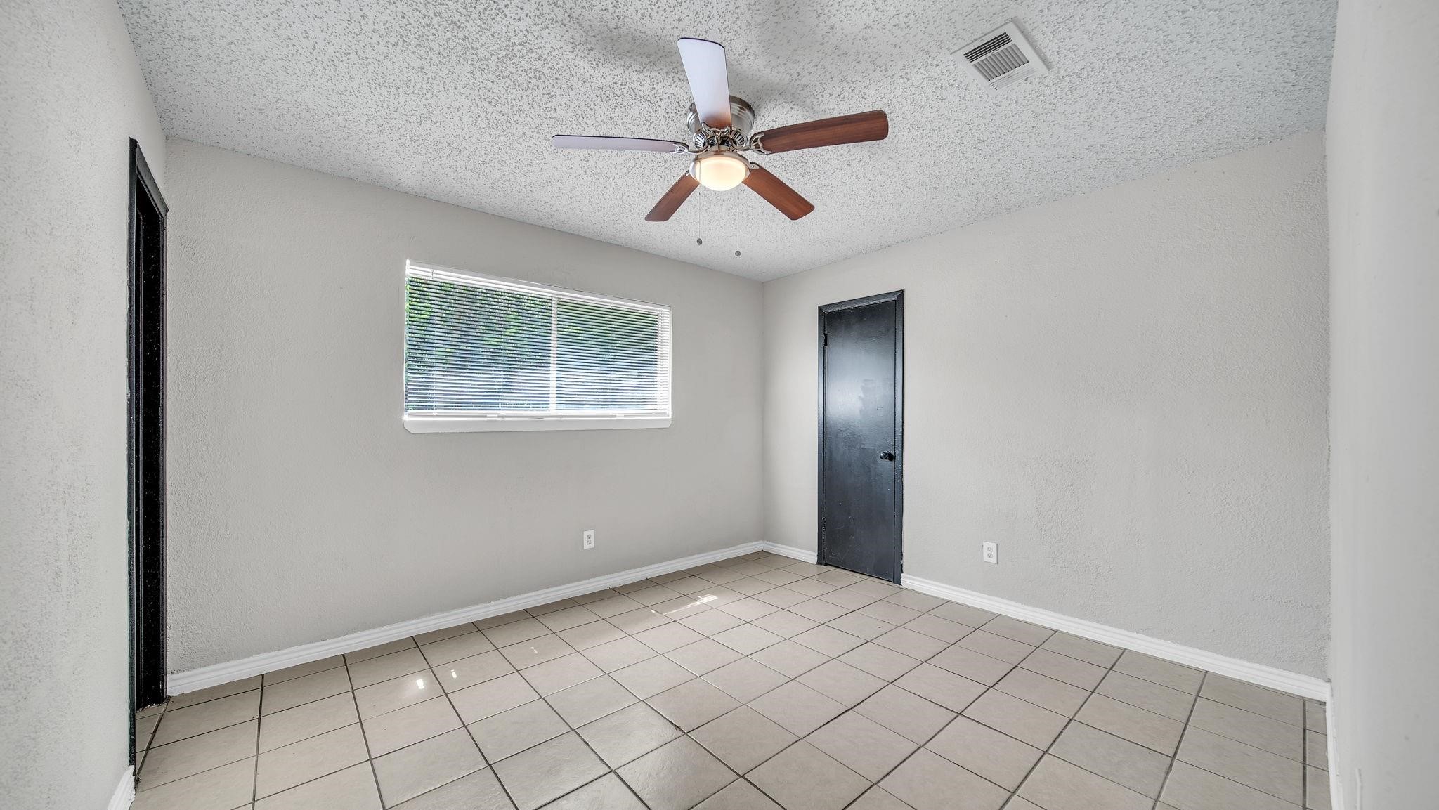 This inviting secondary bedroom features tile flooring, a ceiling fan with integrated lighting, and a spacious walk-in closet. A sliding window overlooks the backyard, welcoming natural light and offering a pleasant view of the outdoor surroundings.