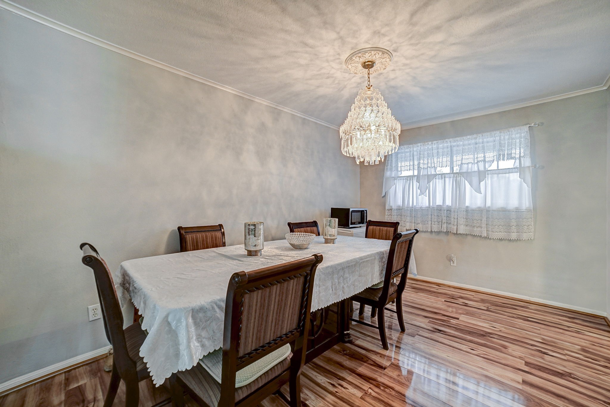 Elegant dining room featuring a stunning chandelier, polished wood flooring, and a large window with lace curtains, creating a bright and inviting space for gatherings.