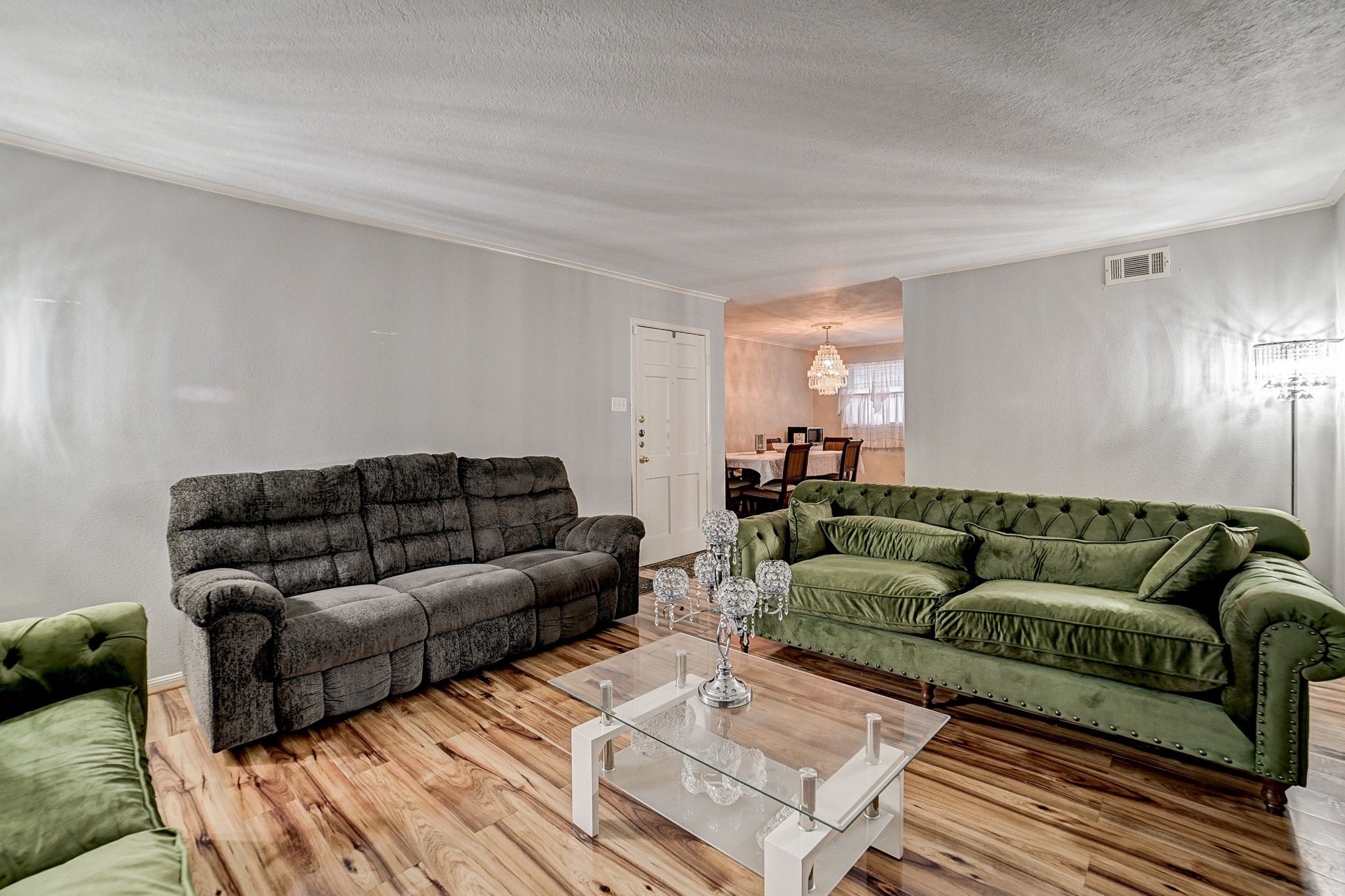 Spacious living room featuring elegant green and gray sofas, a chic glass coffee table, and warm wood flooring, leading into a cozy dining area illuminated by a stunning chandelier.
