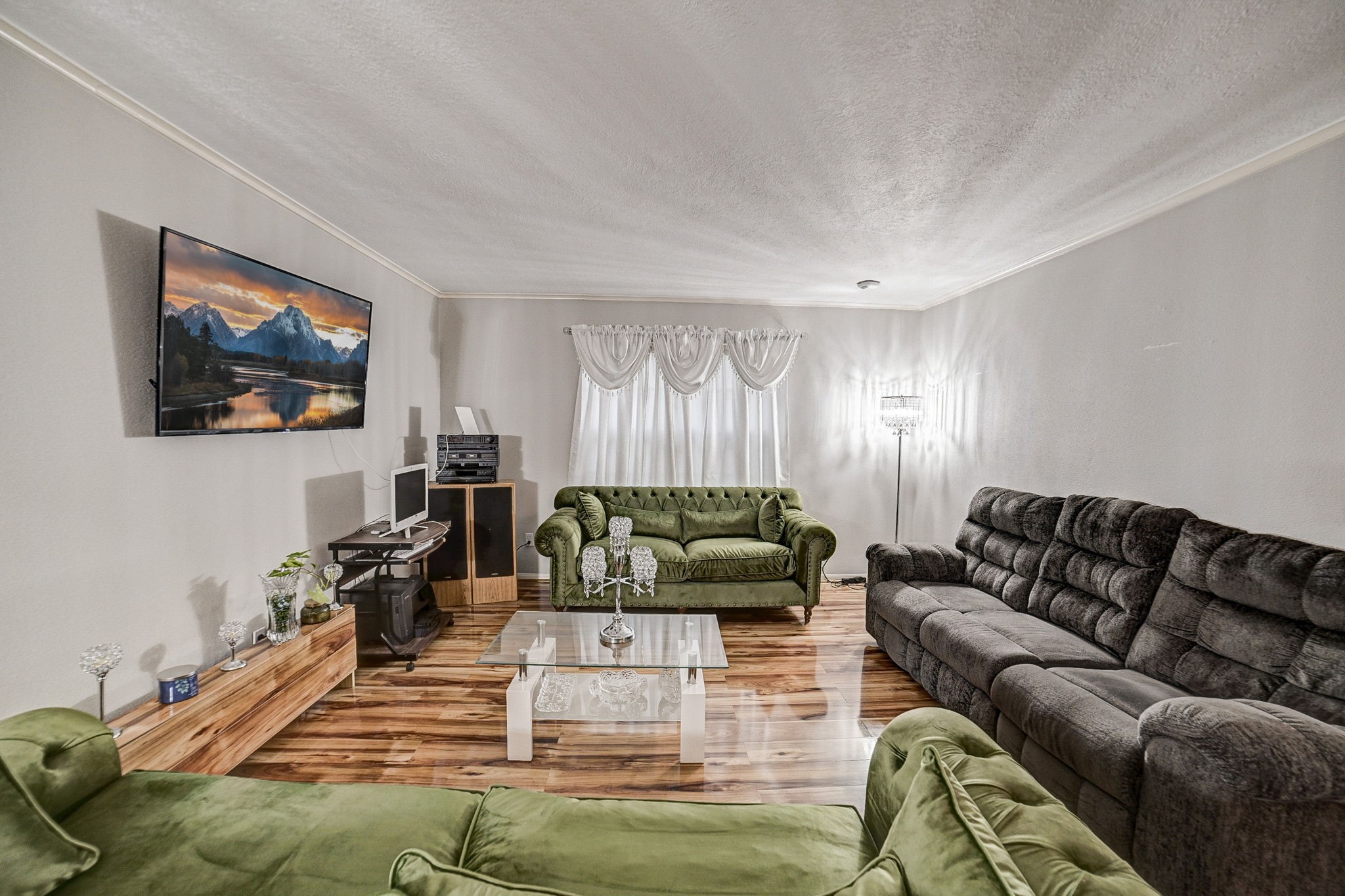 Spacious living room with elegant wood flooring, featuring plush green and gray seating, a wall-mounted TV, and ample natural light from a large window.