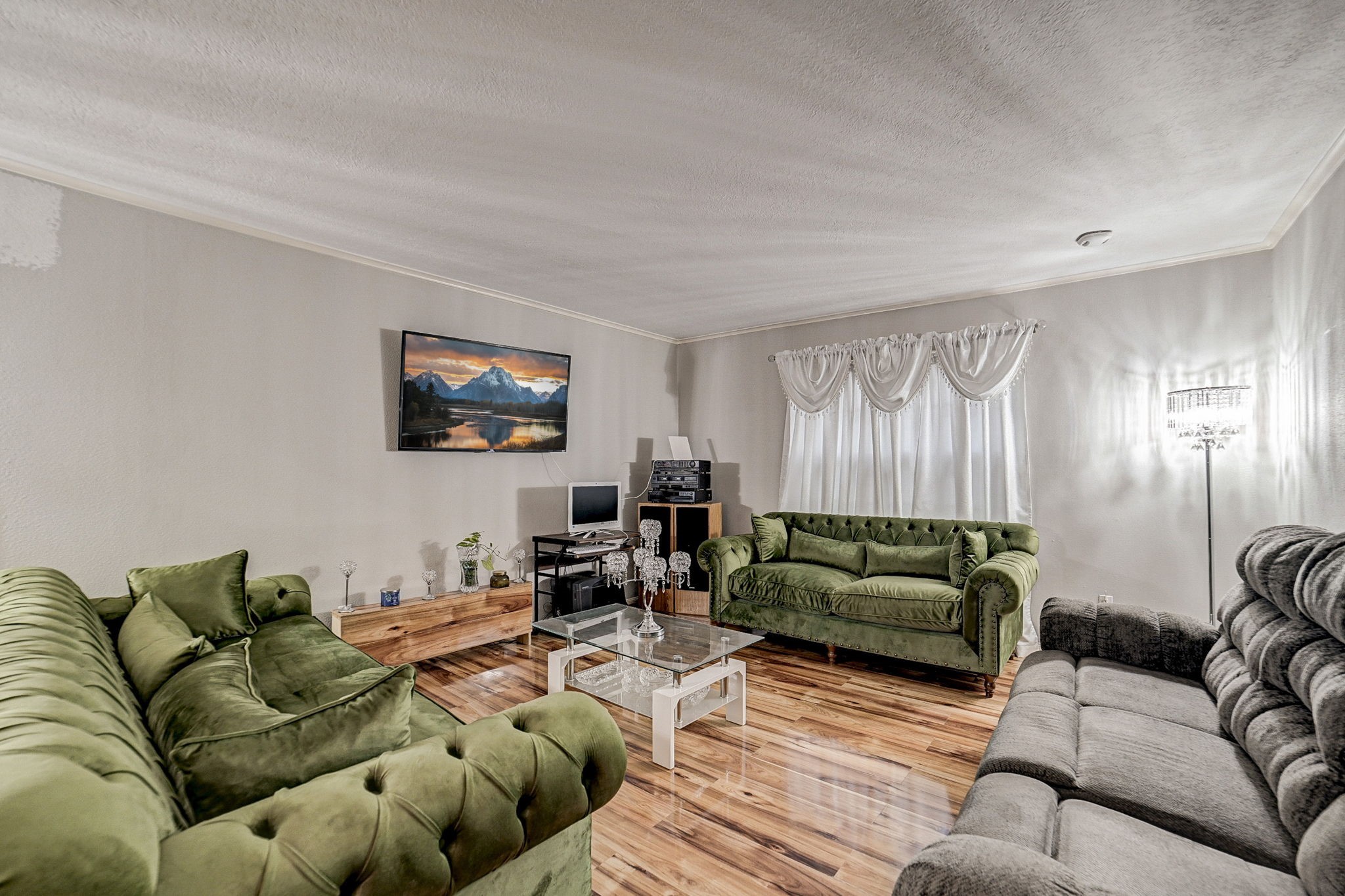Spacious living room featuring elegant green velvet sofas, a large wall-mounted TV, and abundant natural light streaming through stylish drapery. Spacious living room featuring elegant green velvet sofas, a large wall-mounted TV, and abundant natural light streaming through stylish drapery.