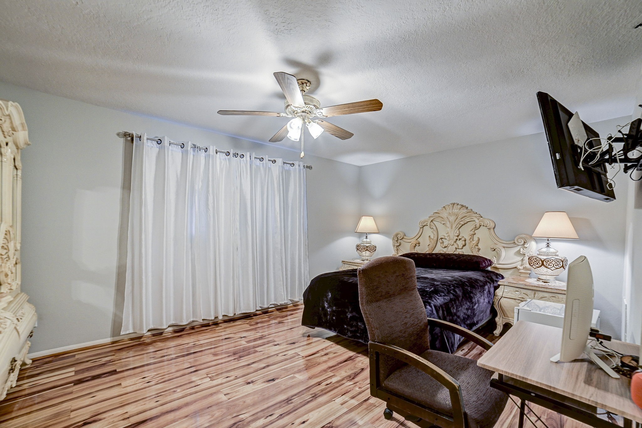 Spacious bedroom featuring elegant wood flooring, a stylish ceiling fan, and a luxurious bed with matching nightstands, complemented by soft lighting and a mounted TV for added convenience.