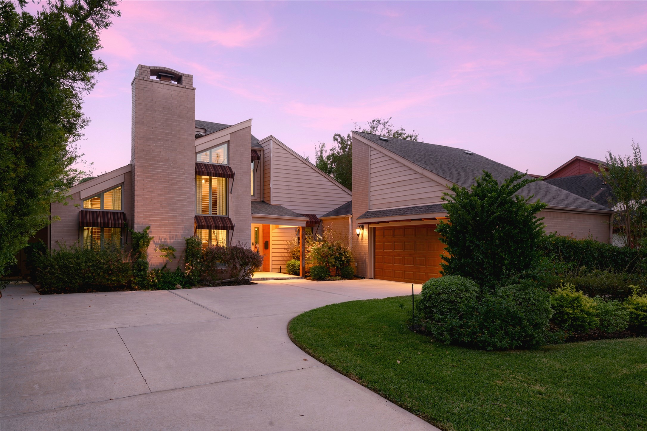 Striking contemporary exterior featuring a tall brick chimney, striped metal awnings & glowing windows at dusk, set behind a sweeping curved driveway & manicured lawn.