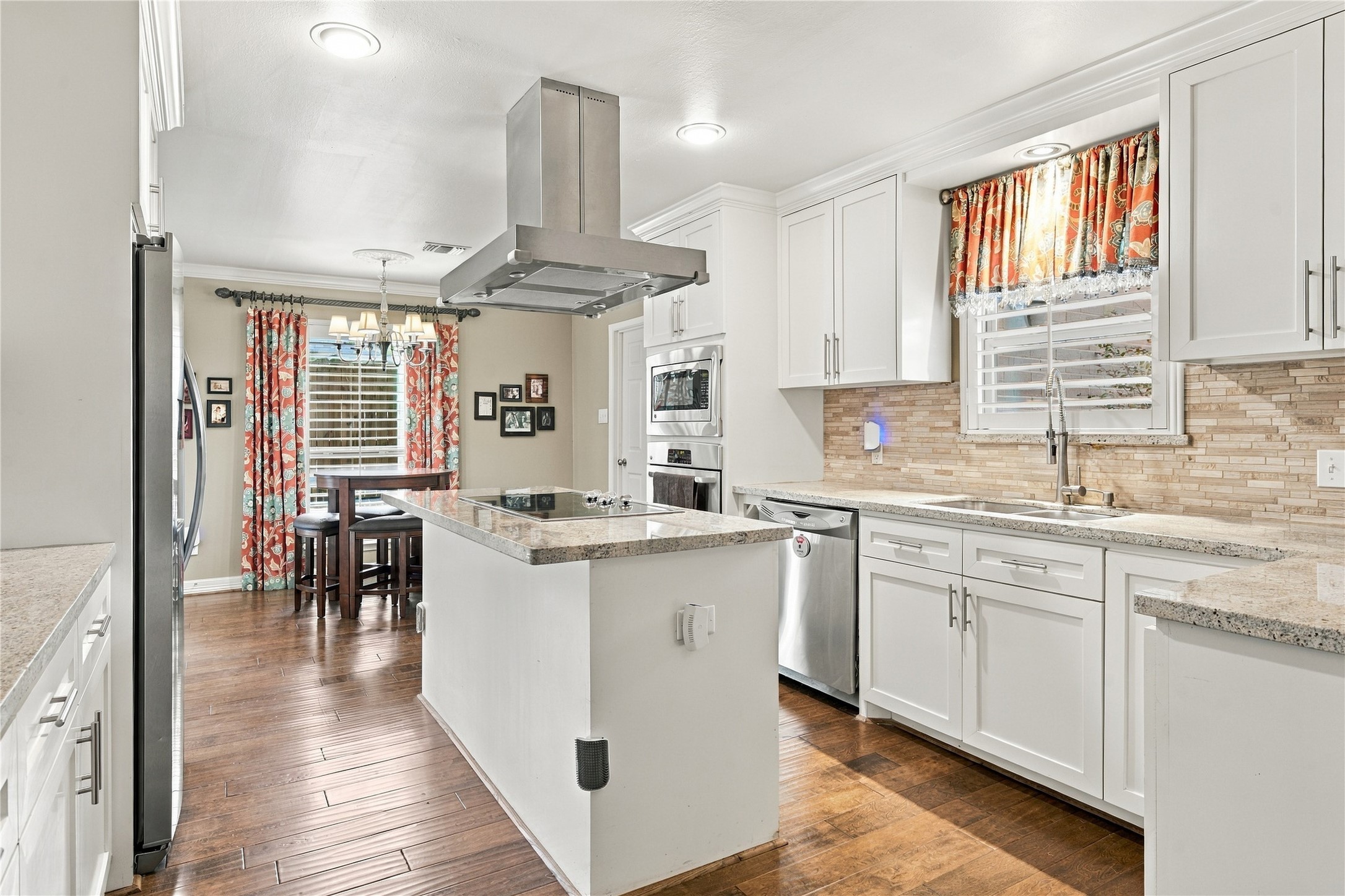 This bright kitchen features white shaker cabinetry, granite countertops, a linear stone mosaic backsplash & stainless steel appliances. The center island houses a smooth-top cooktop beneath a stainless chimney hood, and the wood floors flow into the breakfast room.
