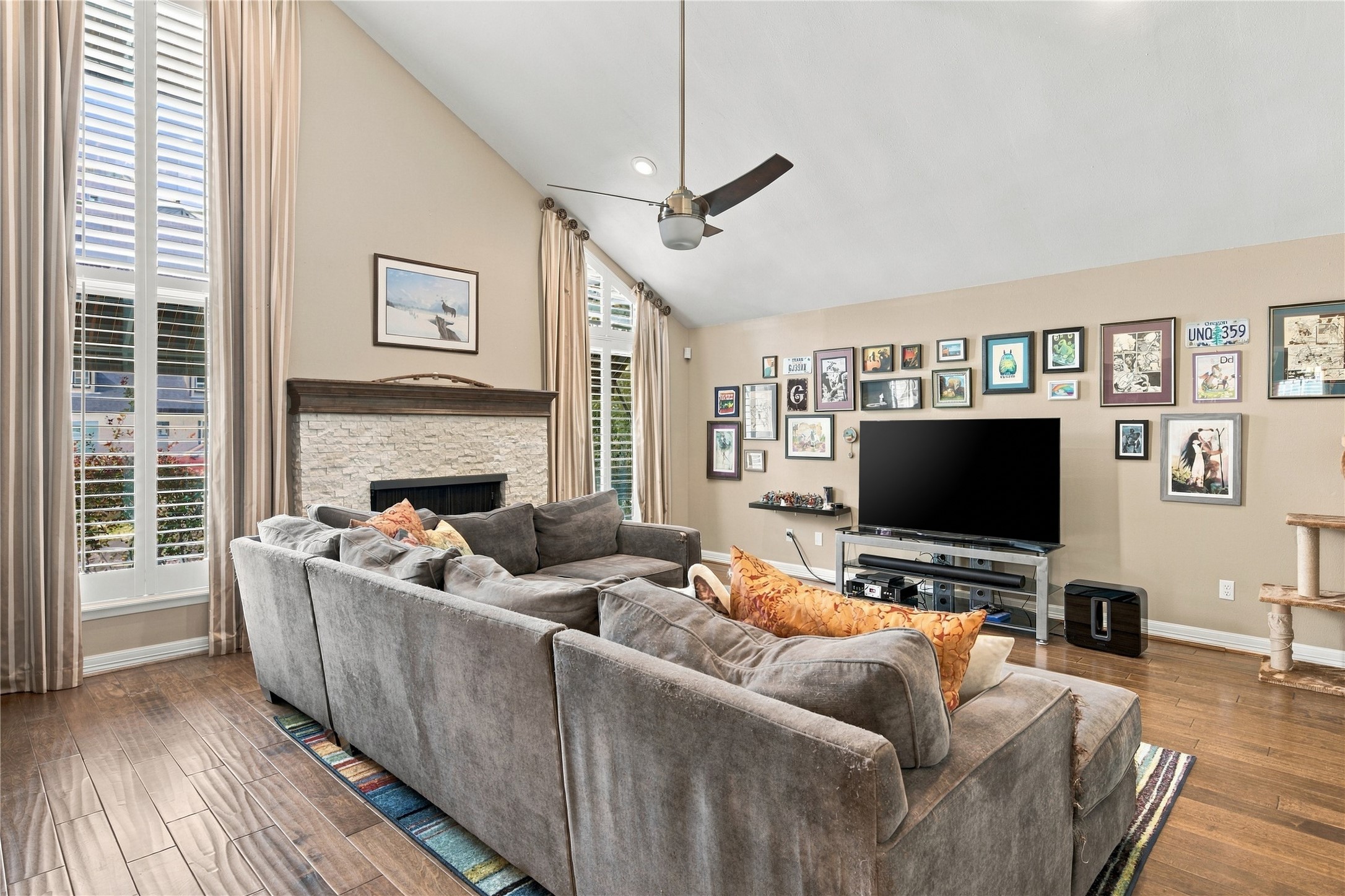 This spacious living room features a stacked-stone gas-log fireplace with dark wood mantel, a dramatic vaulted ceiling & floor-to-ceiling plantation-shuttered windows that fill the space with natural light.
