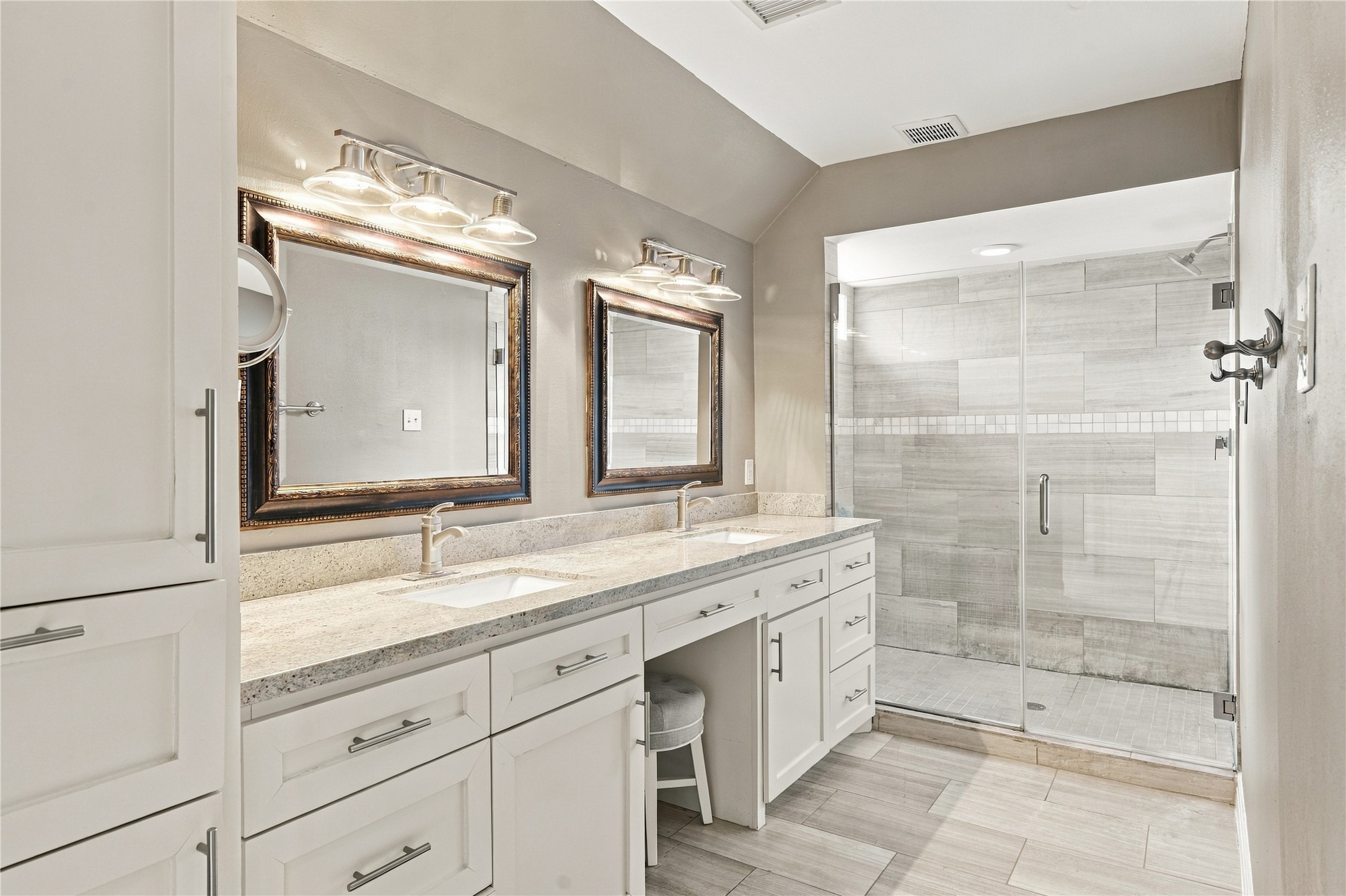 Spa-inspired primary bath features a white shaker dual vanity with granite countertop & built-in makeup station. The oversized frameless glass walk-in shower is wrapped floor to ceiling in large-format gray plank tile with a mosaic accent band.