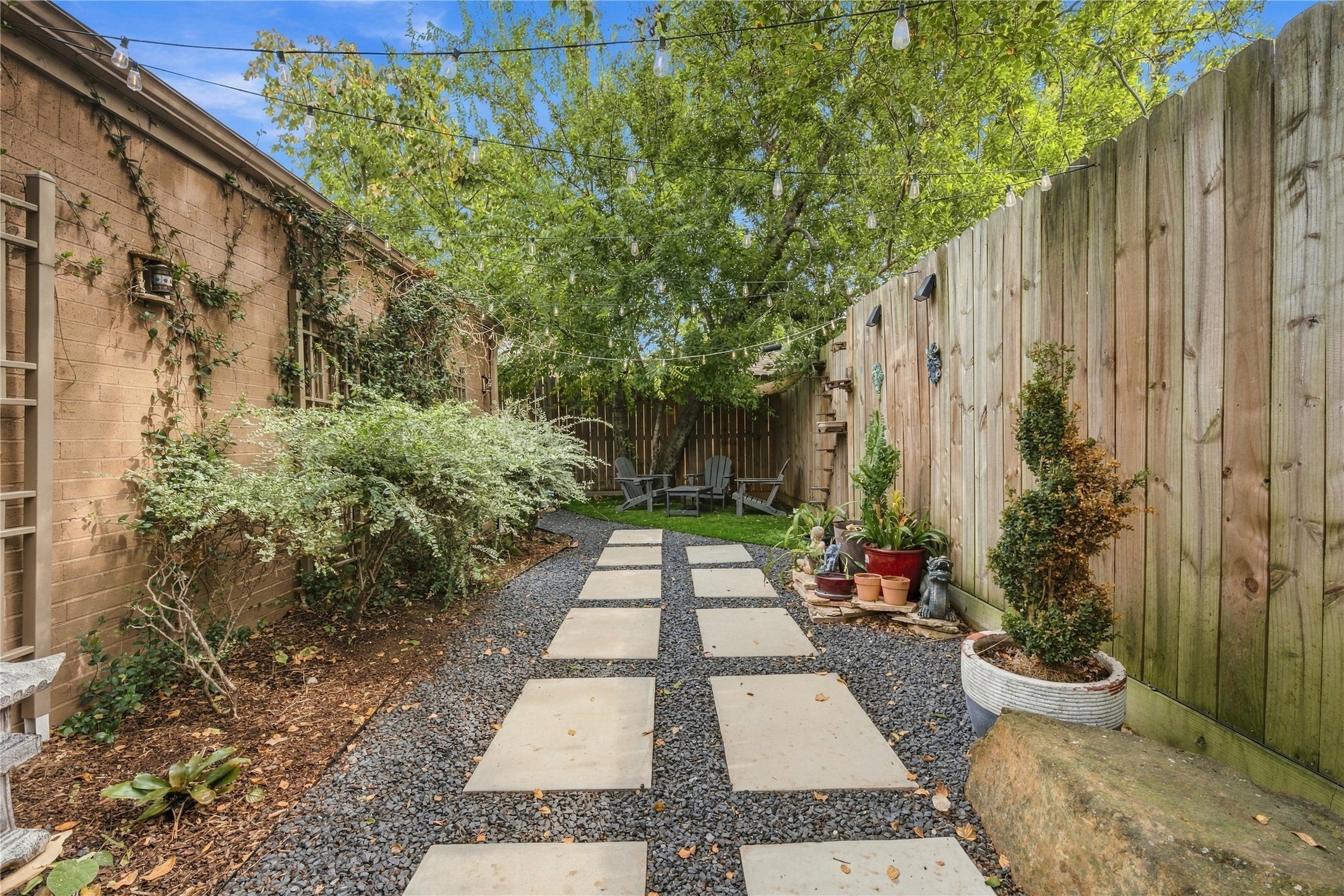 Square stepping stones set in gravel lead down the side yard beneath a canopy of string lights, between a tall wood privacy fence & planted beds along the brick. The path ends at a turf seating area under shaded trees.