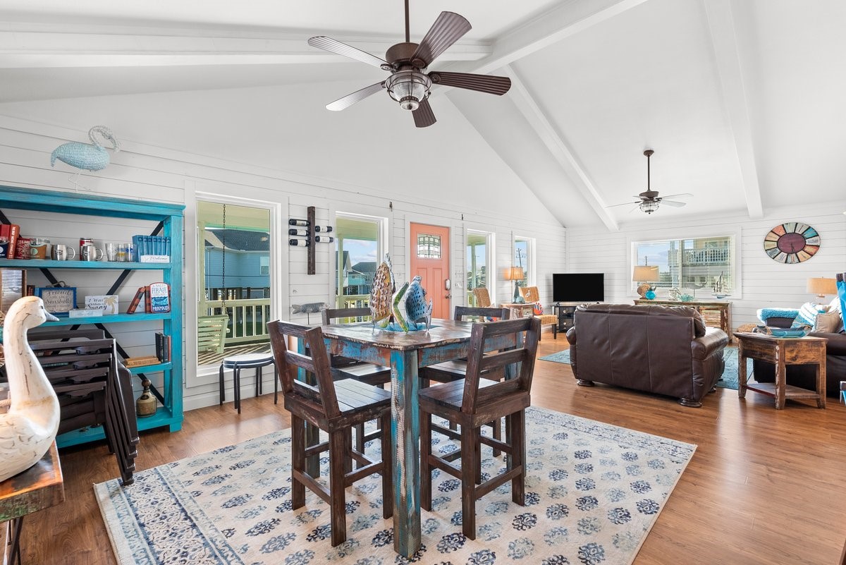 This inviting open-concept living space features high vaulted ceilings, abundant natural light, and stylish wood flooring. The room is tastefully decorated with coastal accents, including a rustic dining set and cozy seating area. Large windows and a charming front door enhance the airy, welcoming atmosphere. This inviting open-concept living space features high vaulted ceilings, abundant natural light, and stylish wood flooring. The room is tastefully decorated with coastal accents, including a rustic dining set and cozy seating area. Large windows and a charming front door enhance the airy, welcoming atmosphere.