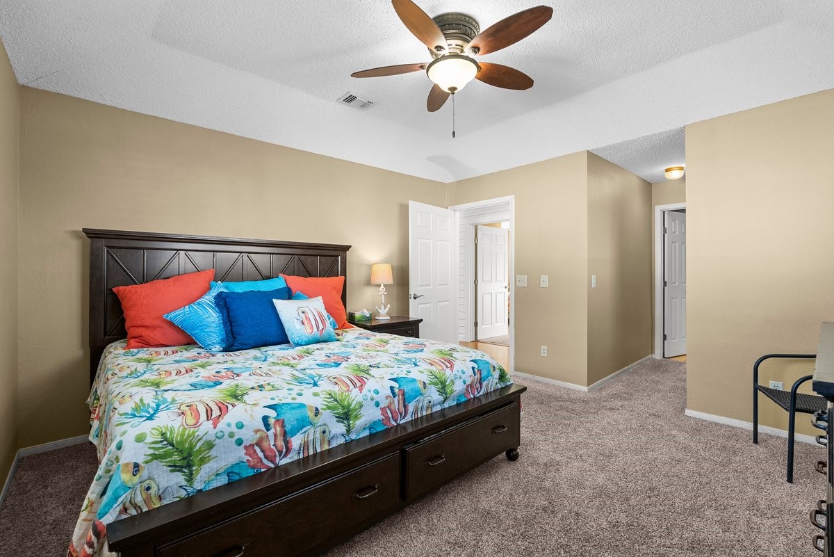 Spacious bedroom with a cozy carpet, featuring a large bed with colorful bedding. The room includes a ceiling fan, neutral walls, and ample natural light, providing a warm and inviting atmosphere.