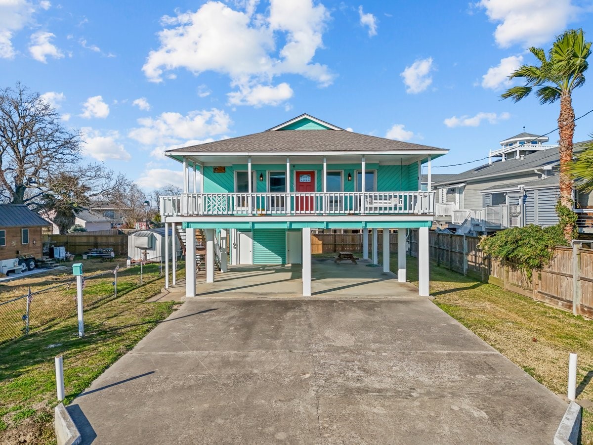 Elevated coastal home with a vibrant exterior, featuring a spacious driveway and covered parking. A wraparound porch provides charm and outdoor living space, while a fenced yard offers privacy. Perfect for enjoying seaside living.