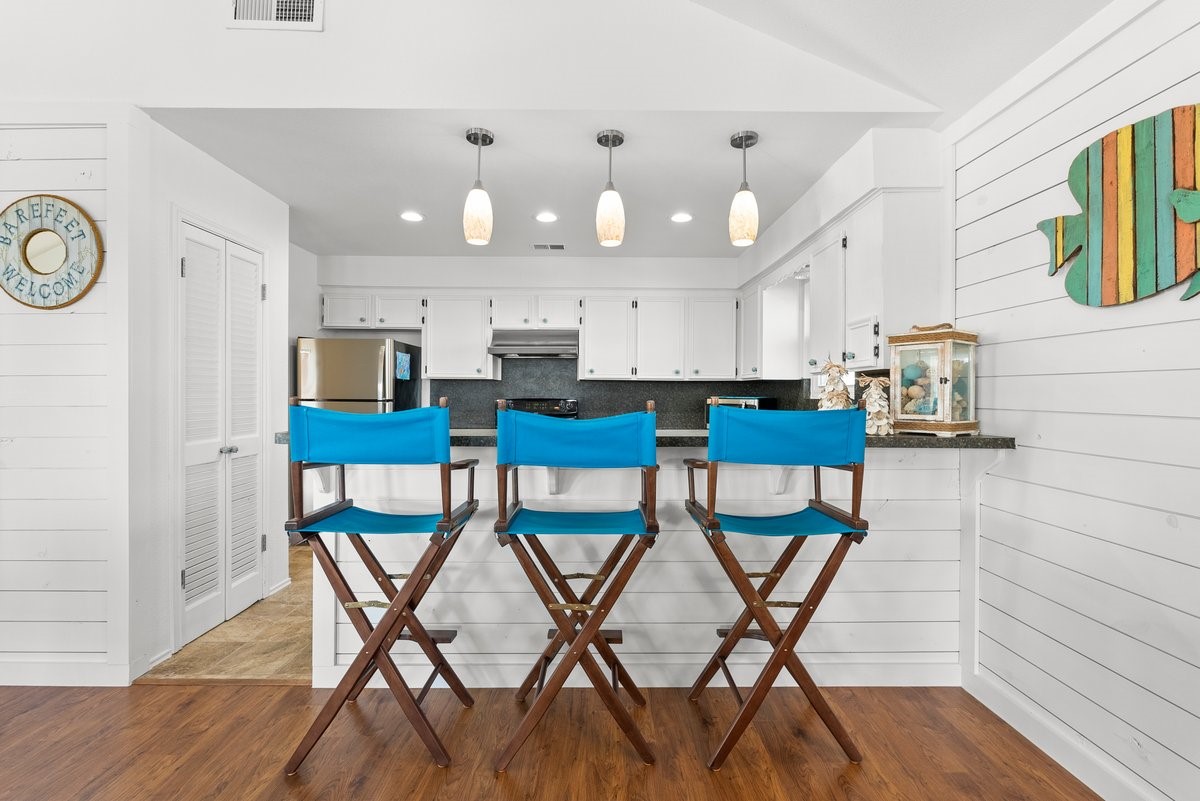 Bright, coastal-style kitchen with white cabinets and shiplap walls. Features a breakfast bar with three blue director's chairs, pendant lighting, and stainless steel appliances.
