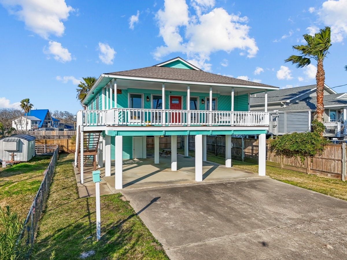 This elevated coastal home features a vibrant turquoise exterior with a spacious wraparound porch, covered parking, and a fenced yard. Ideal for enjoying beachside living.