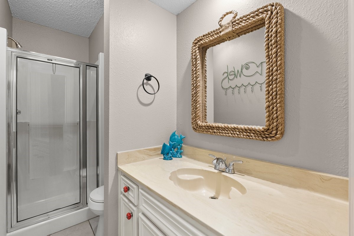 This bathroom features a clean, modern design with a glass-enclosed shower, a beige countertop with a sink, and a decorative rope-framed mirror. The neutral tones and simple decor create a welcoming, functional space.