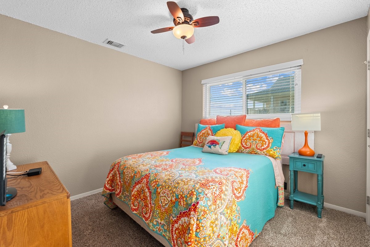 Cozy bedroom with neutral walls and carpet, featuring a colorful bedspread and pillows. It has a ceiling fan, a large window for natural light, and a mix of wooden and painted furniture for a vibrant touch.