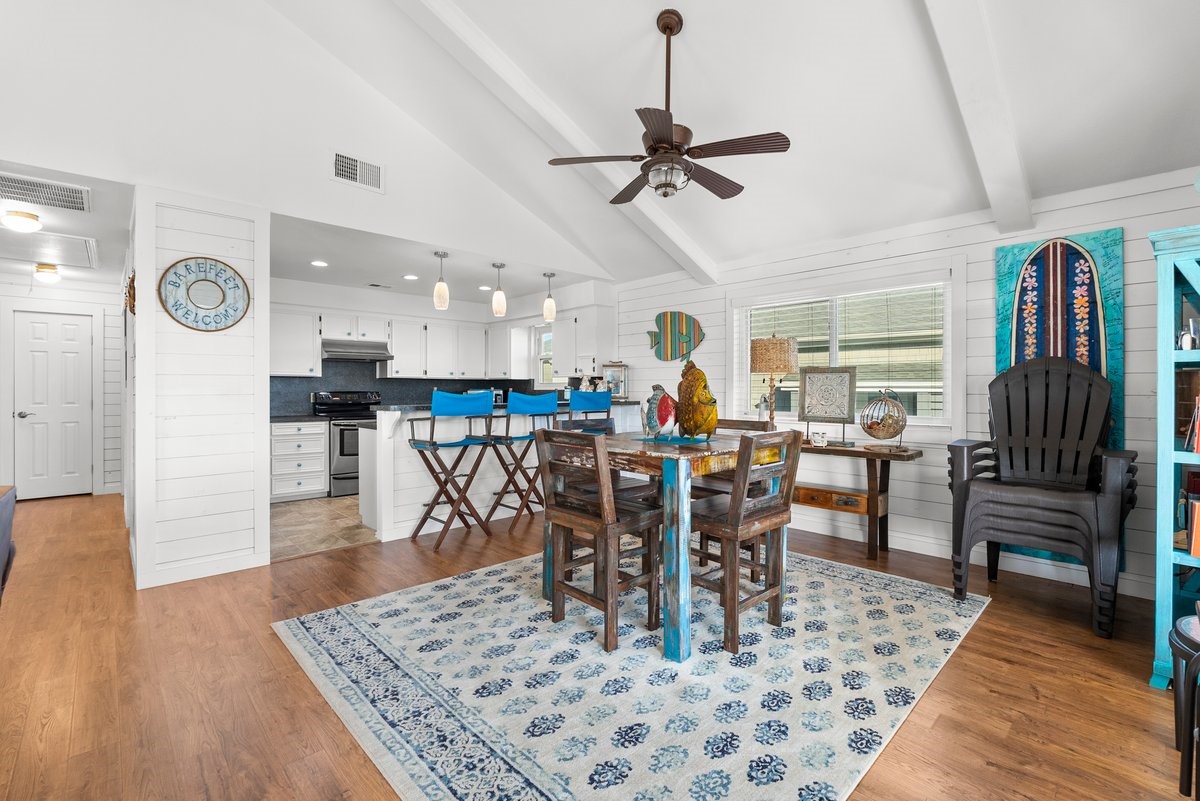 This bright, open-concept living space features a vaulted ceiling and hardwood floors. The kitchen has white cabinets, stainless steel appliances, and a breakfast bar with blue stools. The dining area is cozy, with colorful decor creating a welcoming atmosphere.