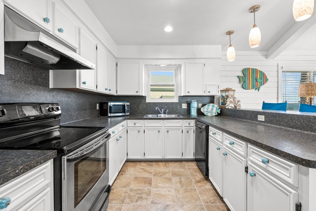 This kitchen features a bright, coastal design with white cabinetry, dark countertops, and stainless steel appliances. It includes a spacious layout with ample counter space, modern light fixtures, and decorative accents, creating a welcoming and functional cooking area.