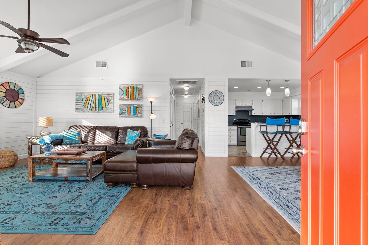 This open-concept living space features high vaulted ceilings and wood flooring, creating a spacious feel. The room is bright and welcoming with a pop of color from the orange door.