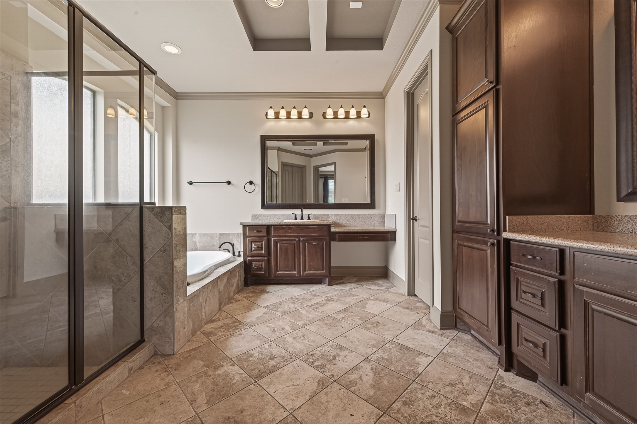 The spacious primary bathroom features a tray ceiling with crown molding, tile flooring, and rich wood cabinetry with granite countertops. A glass enclosed shower and a soaking tub provide the perfect blend of luxury and practicality. A tall linen cabinet on the right offers generous storage, keeping the space organized and clutter free.