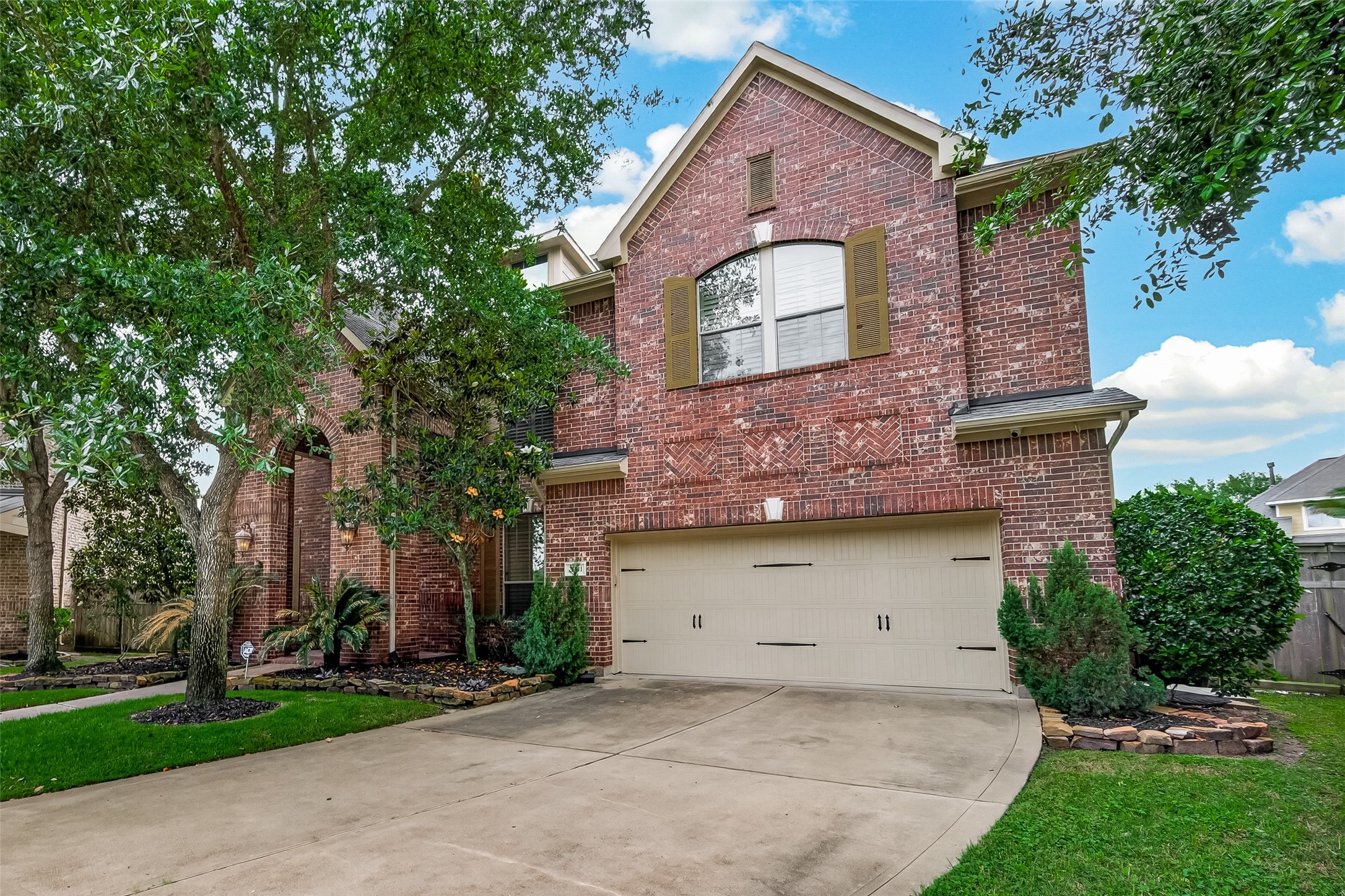 From this angle, the home's rich brick exterior shines, showcasing the decorative herringbone brickwork detail and arched upper window with classic shutters. Mature trees frame the property beautifully, adding depth and natural charm to the streetscape. The wide driveway leads to the two-car attached garage, while neatly edged plant beds with evergreen shrubs and tropical foliage enhance the home's polished and inviting curb appeal.