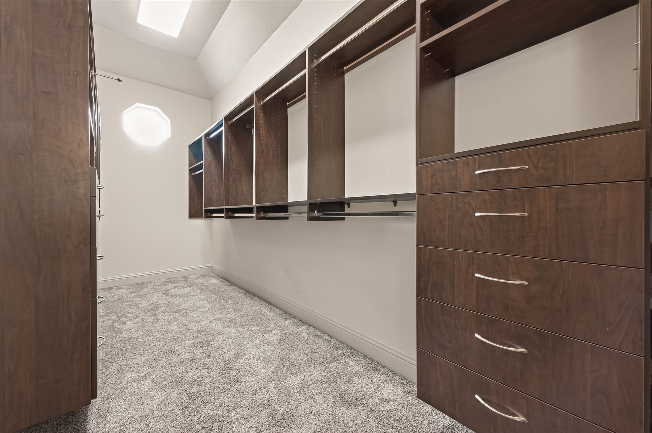 The spacious full entry closet features a custom built in organizer system with rich wood finish, including multiple hanging rods, adjustable shelving, and drawers for optimal storage. Plush carpeting adds comfort underfoot, while a skylight and an octagonal window bring in natural light. This well designed closet makes it easy to keep your wardrobe neatly organized and accessible.