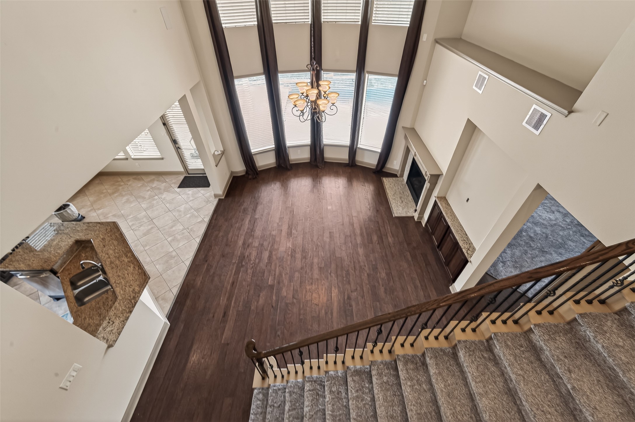 From the upper level, this breathtaking view captures the grandeur of the home's open layout. Soaring two-story ceilings, a dramatic curved wall of windows, and an elegant chandelier create a striking focal point in the living area below. Rich hardwood flooring anchors the space, while a fireplace with a granite surround adds warmth and sophistication. The staircase with its wooden handrail and wrought iron balusters leads gracefully to this impressive vantage point, with the kitchen visible just beyond.