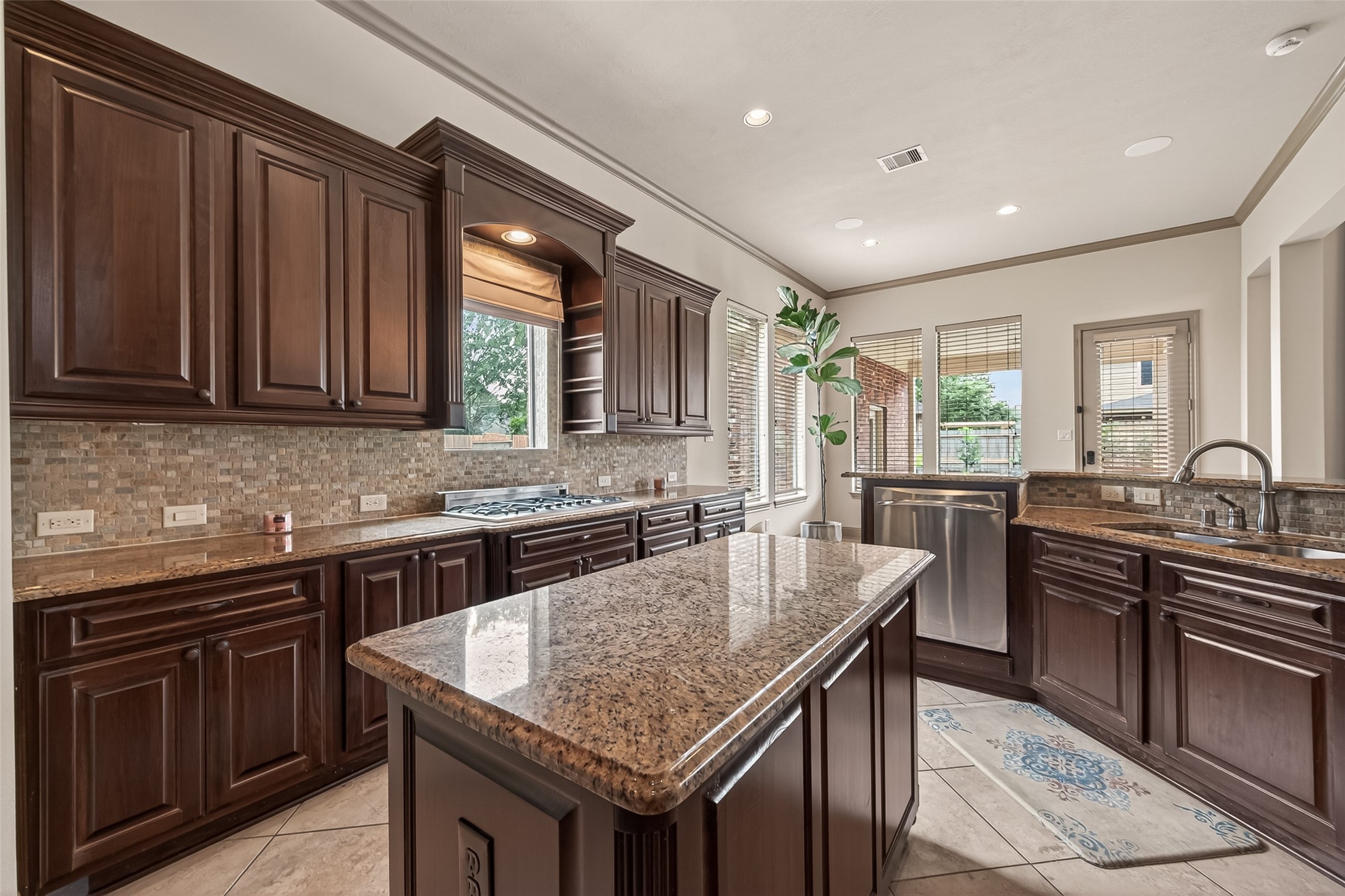 From this angle, the kitchen's thoughtful layout truly shines. Rich dark wood cabinetry lines the walls, complemented by granite countertops and a stone mosaic backsplash. The gas cooktop is perfectly positioned beneath a window that fills the space with natural light, while multiple windows along the back wall keep the kitchen bright and airy. The central island offers generous prep space, and the stainless steel dishwasher adds everyday convenience. A dual-sink setup ensures effortless meal prep and clean