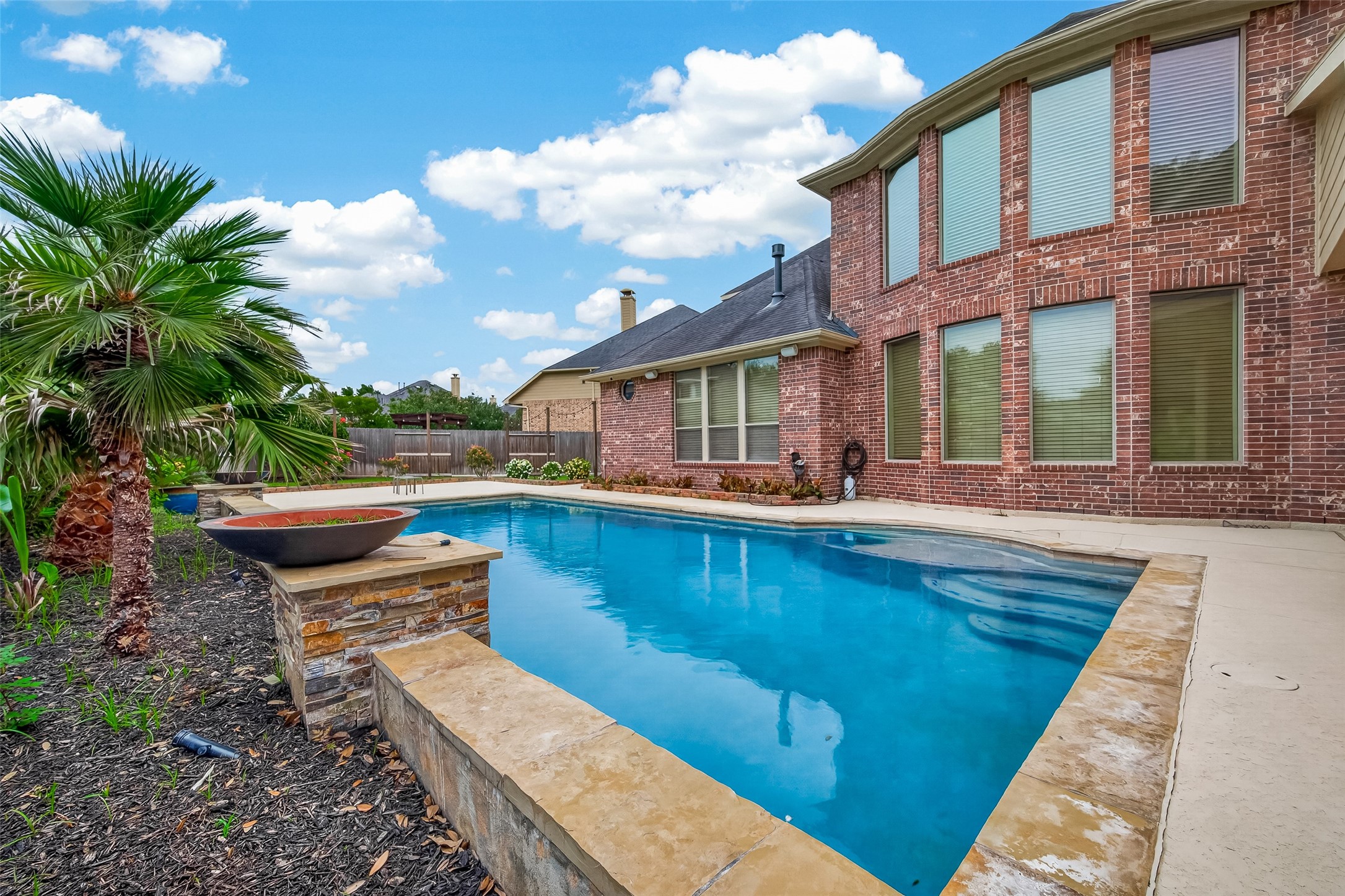This view from poolside showcases the stunning combination of the sparkling pool and the home's impressive brick rear elevation. A raised stone planter adds architectural interest to the pool area, while lush tropical palms and flowering plants create a vibrant, private backdrop. The covered patio just off the home provides a shaded retreat, making this backyard a perfect setting for entertaining loved ones or simply unwinding on a beautiful day.