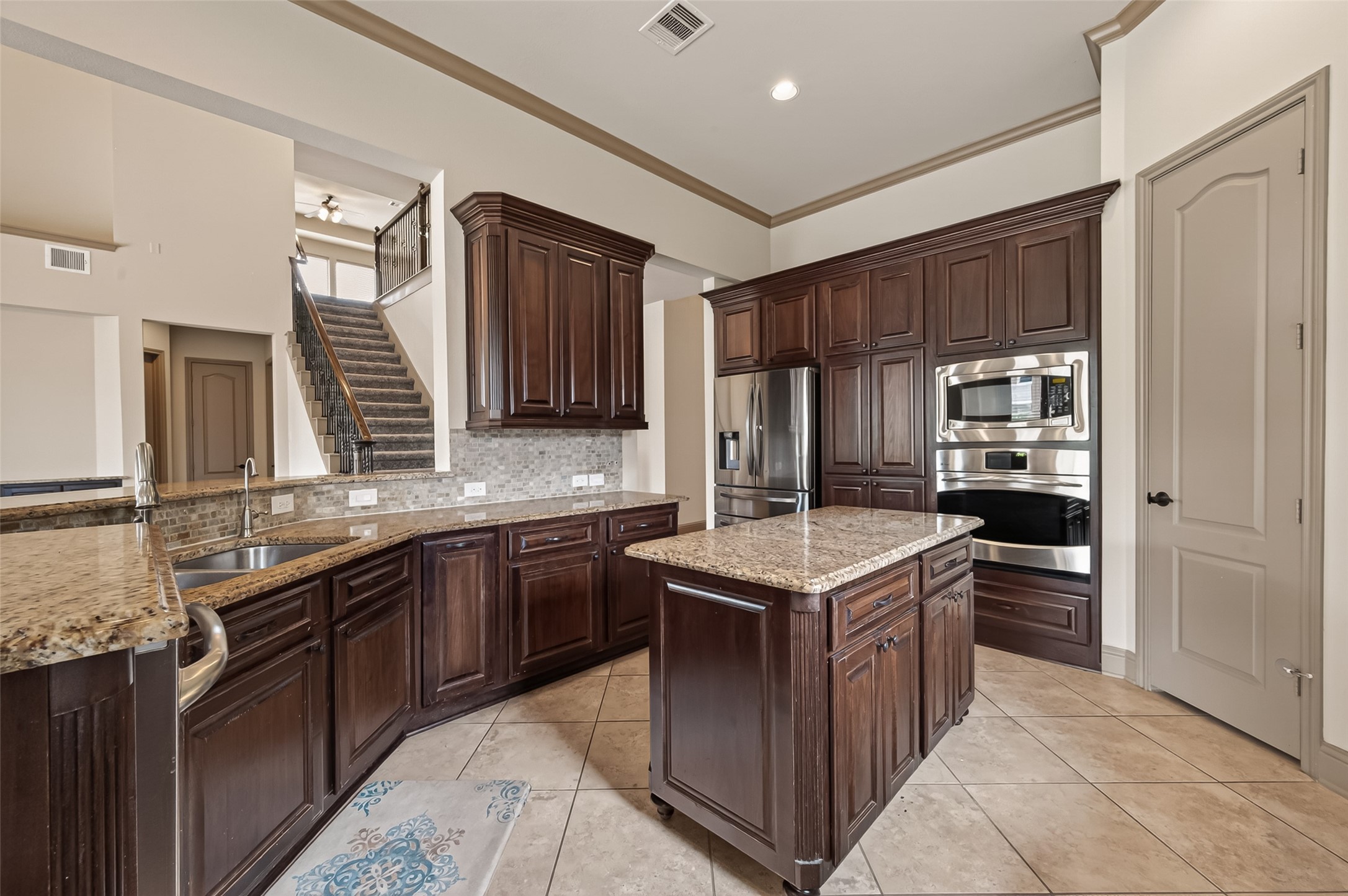 This expansive gourmet kitchen is a chef's dream, featuring rich dark wood cabinetry with raised panel detailing, granite countertops, and a tasteful mosaic tile backsplash. Stainless steel appliances include a built-in double oven, microwave, and refrigerator with water dispenser. A central island provides additional prep space and storage, while tile flooring and crown molding add a refined finishing touch. From this vantage point, the elegant staircase with wrought iron railing is visible, showcasing the