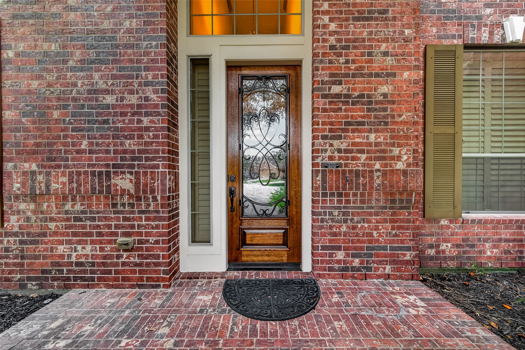 A stunning solid wood front door with intricate wrought iron scrollwork and a glass panel makes a grand first impression. Flanked by a sidelite and topped with a transom window that glows with warm light, this entryway perfectly balances elegance and charm. The classic brick porch floor adds a timeless finishing touch, welcoming you and your guests in style.