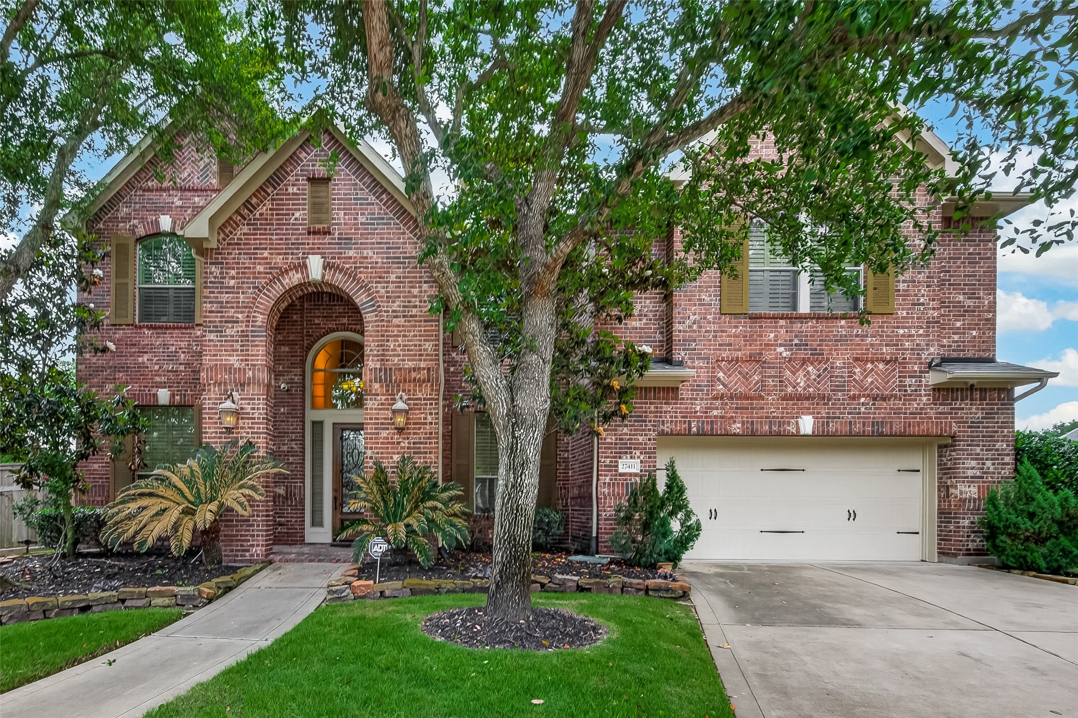 This stunning two-story brick home makes a lasting first impression with its classic red brick facade, decorative herringbone brickwork accents, and a beautiful arched entryway framed by coach lantern lighting. A mature tree anchors the front lawn, adding natural shade and character to the lush, well-maintained landscaping. The two-car attached garage and wide driveway provide ample parking, while the thoughtfully designed plant beds with tropical foliage complete this home's gorgeous curb appeal.