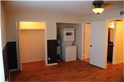 This photo shows a room with hardwood flooring, a ceiling light, and a compact laundry area with a stacked washer and dryer. There are two open doorways leading to a closet and another room.