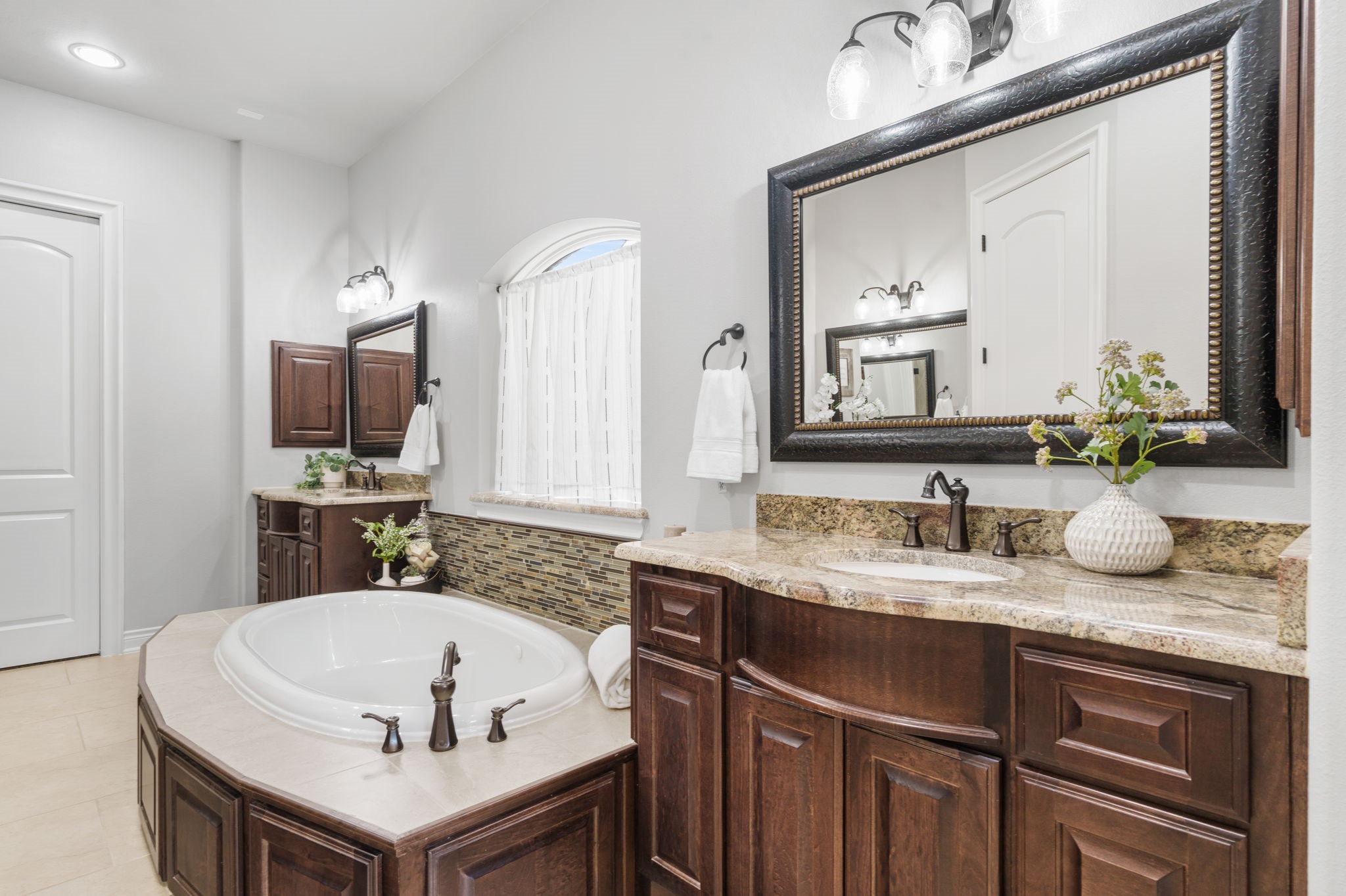 Spa-inspired primary bath featuring separate granite-topped vanities, rich cabinetry, and a spacious soaking tub.