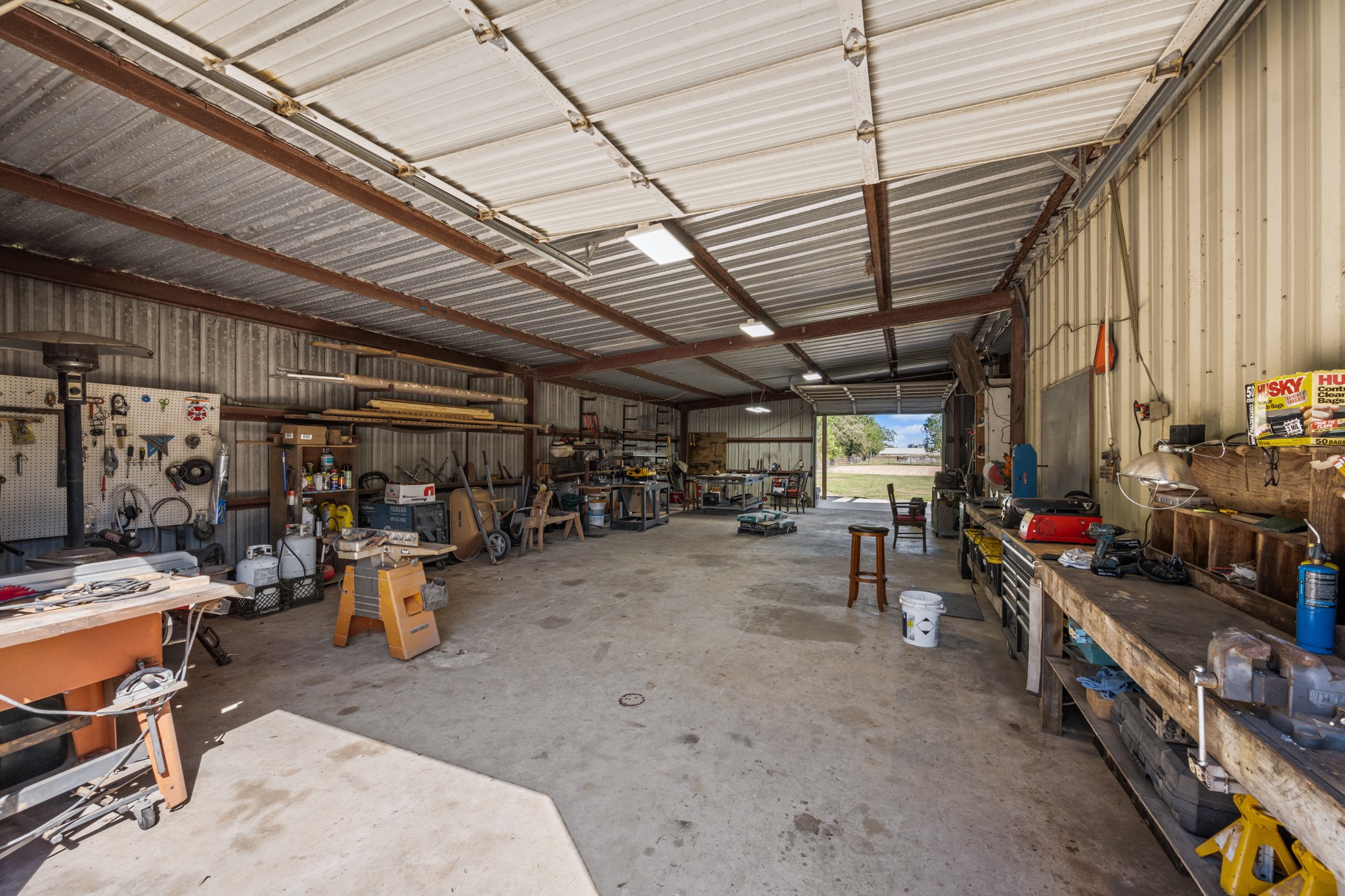 Expansive metal workshop featuring concrete flooring, overhead lighting, exposed steel beams, multiple workbenches, and generous space for tools, equipment, and projects.