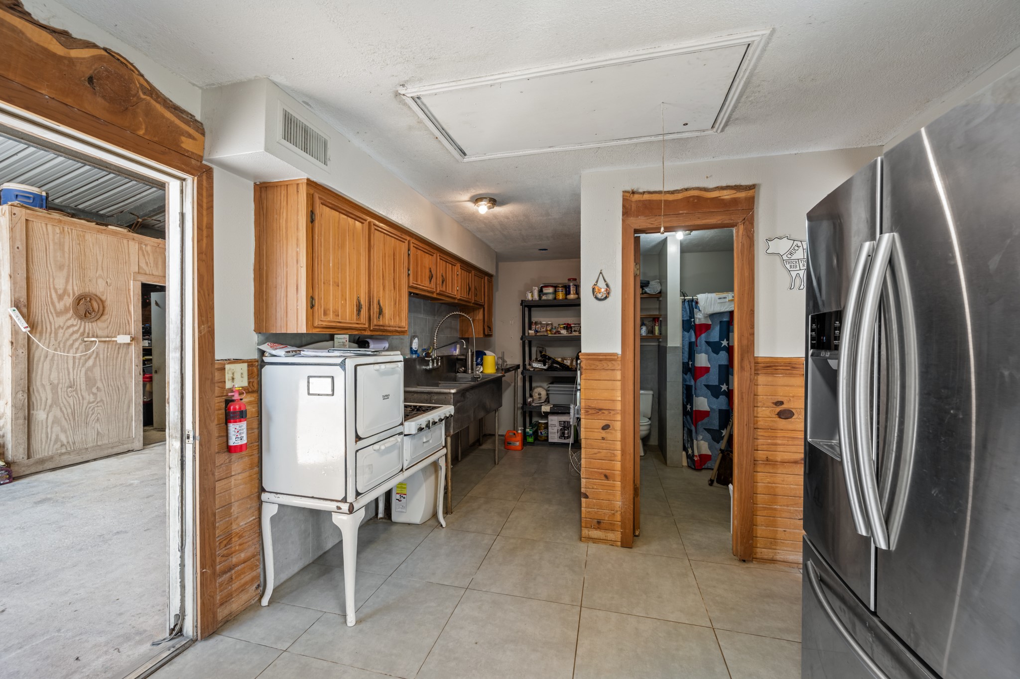 Convenient secondary kitchen and utility space featuring wood cabinetry, stainless steel sink and work surfaces, tiled flooring, refrigeration, and an adjoining bathroom.