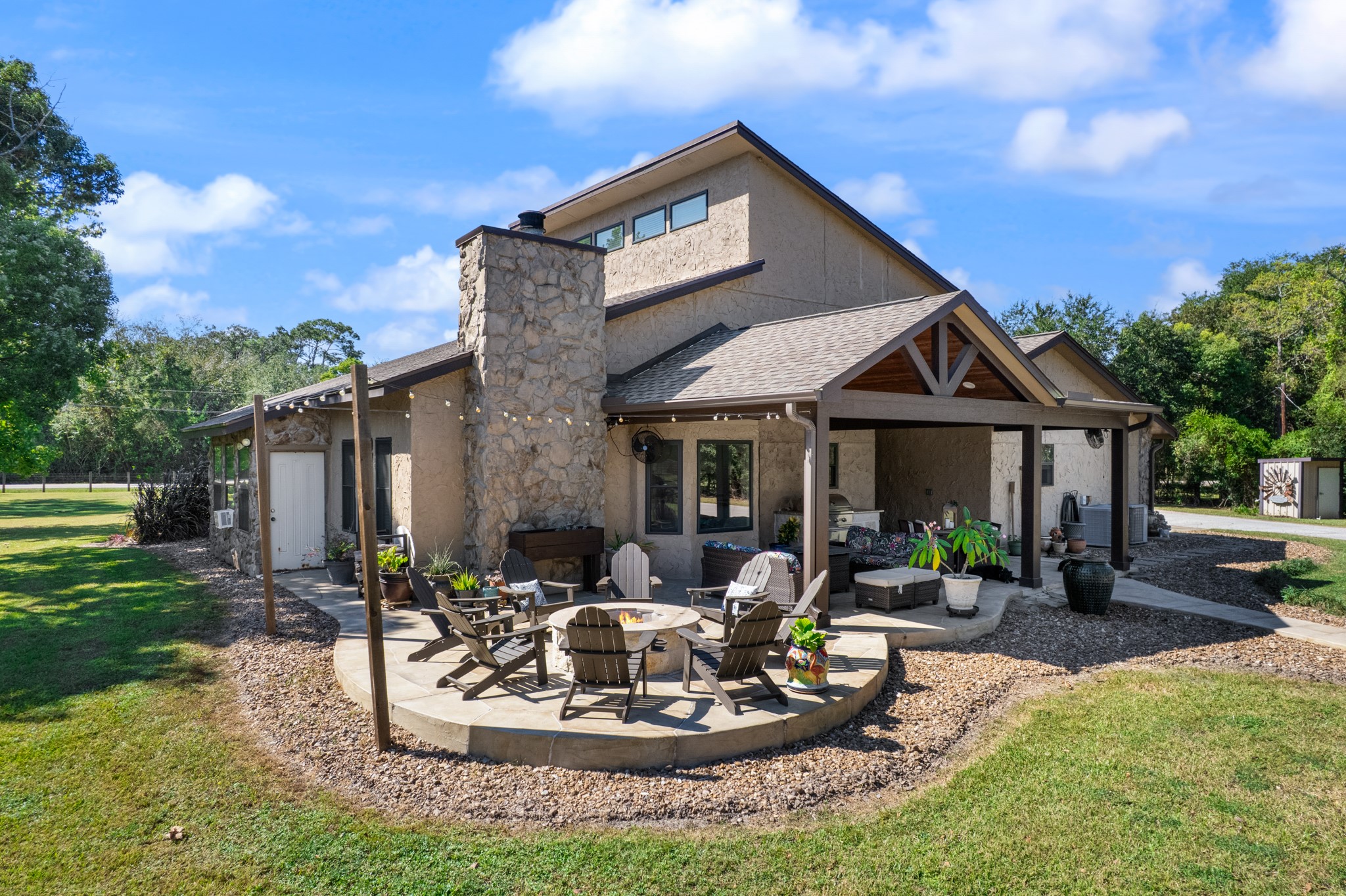 Designed for entertaining, this expansive backyard retreat features a covered outdoor living and dining area, stone fireplace, and circular patio with a fire pit surrounded by Adirondack seating. Beautiful stone accents and mature trees complete this inviting outdoor setting.